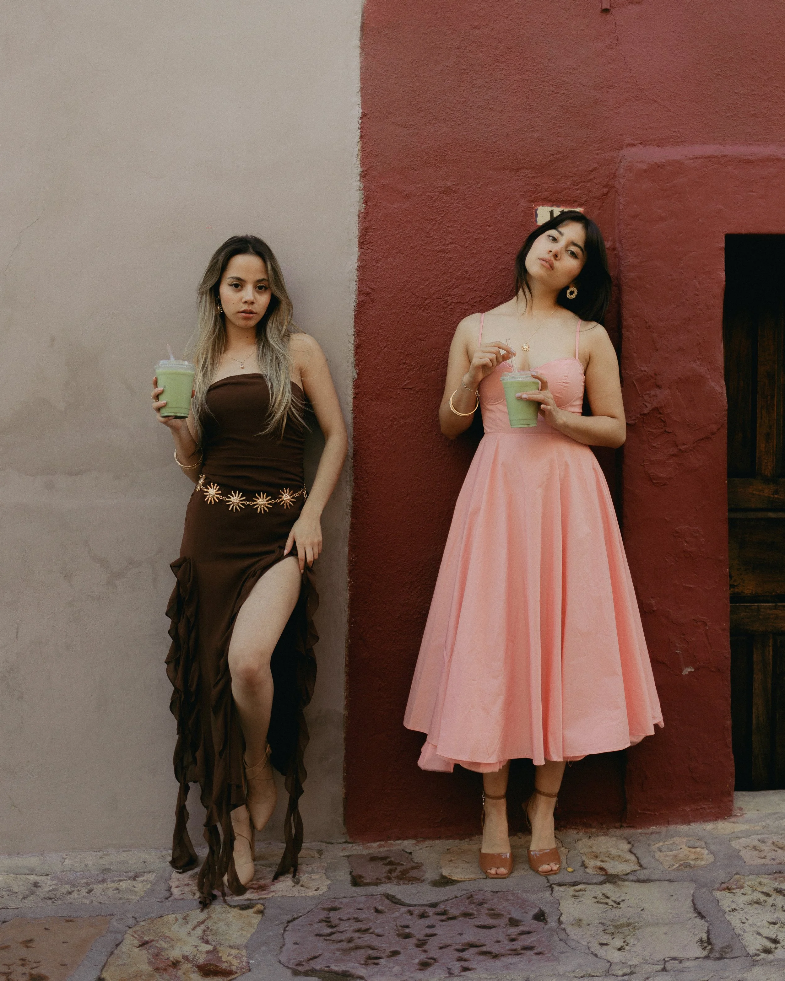 Two young women standing against a wall, holding green drinks with straws, dressed in stylish dresses, one in a brown dress with embroidery and high slit, the other in a pink gown with spaghetti straps, both wearing heels and jewelry.