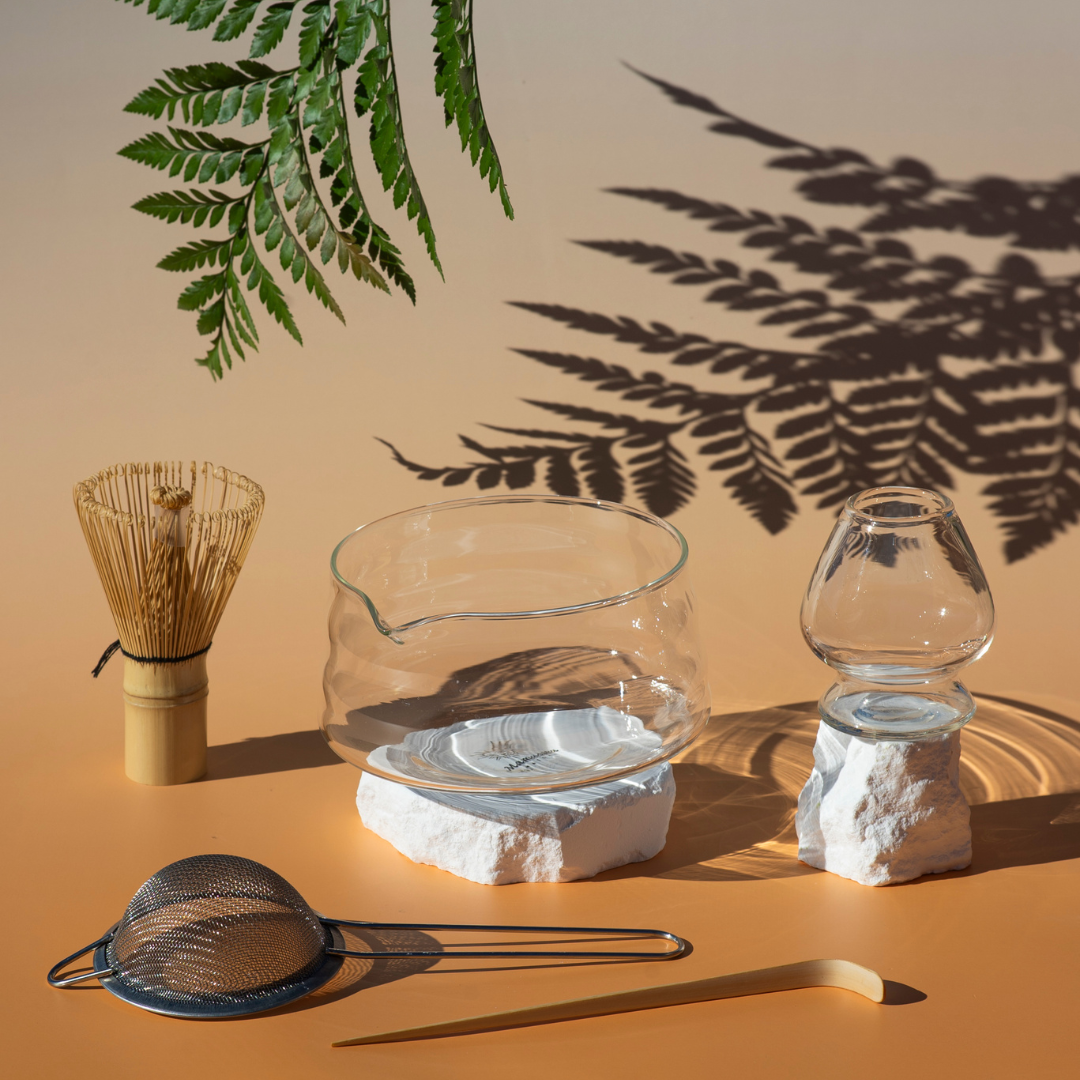 Glass bowl and glass vase on white rocks, bamboo whisk, metal strainer, and wooden chopstick on a peach-colored surface, with green fern leaves casting shadows on a beige background.