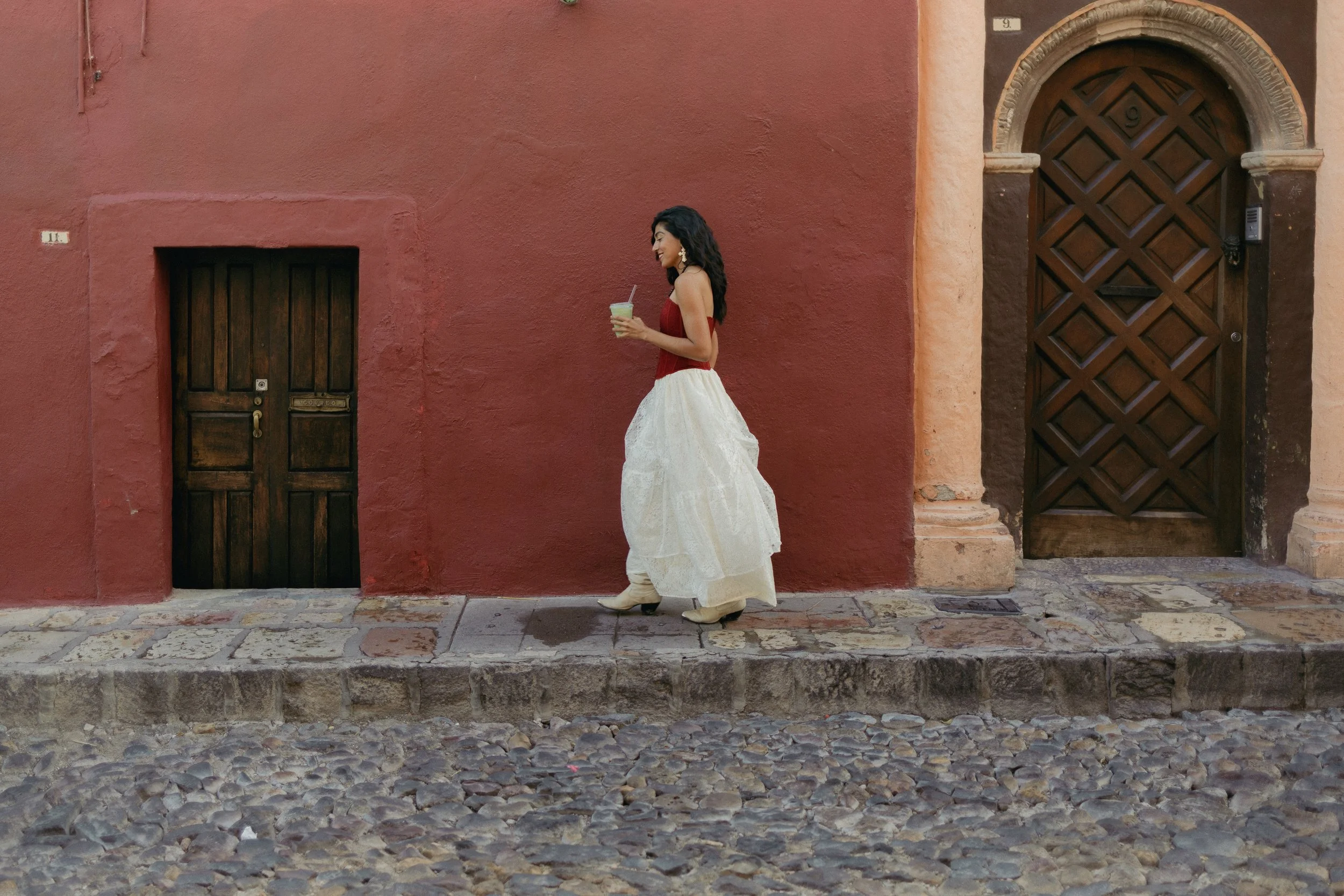 A woman in a white dress and red top walking on a cobblestone street while holding a cold beverage in a plastic cup with a straw, in front of a red and peach building with dark wooden doors.