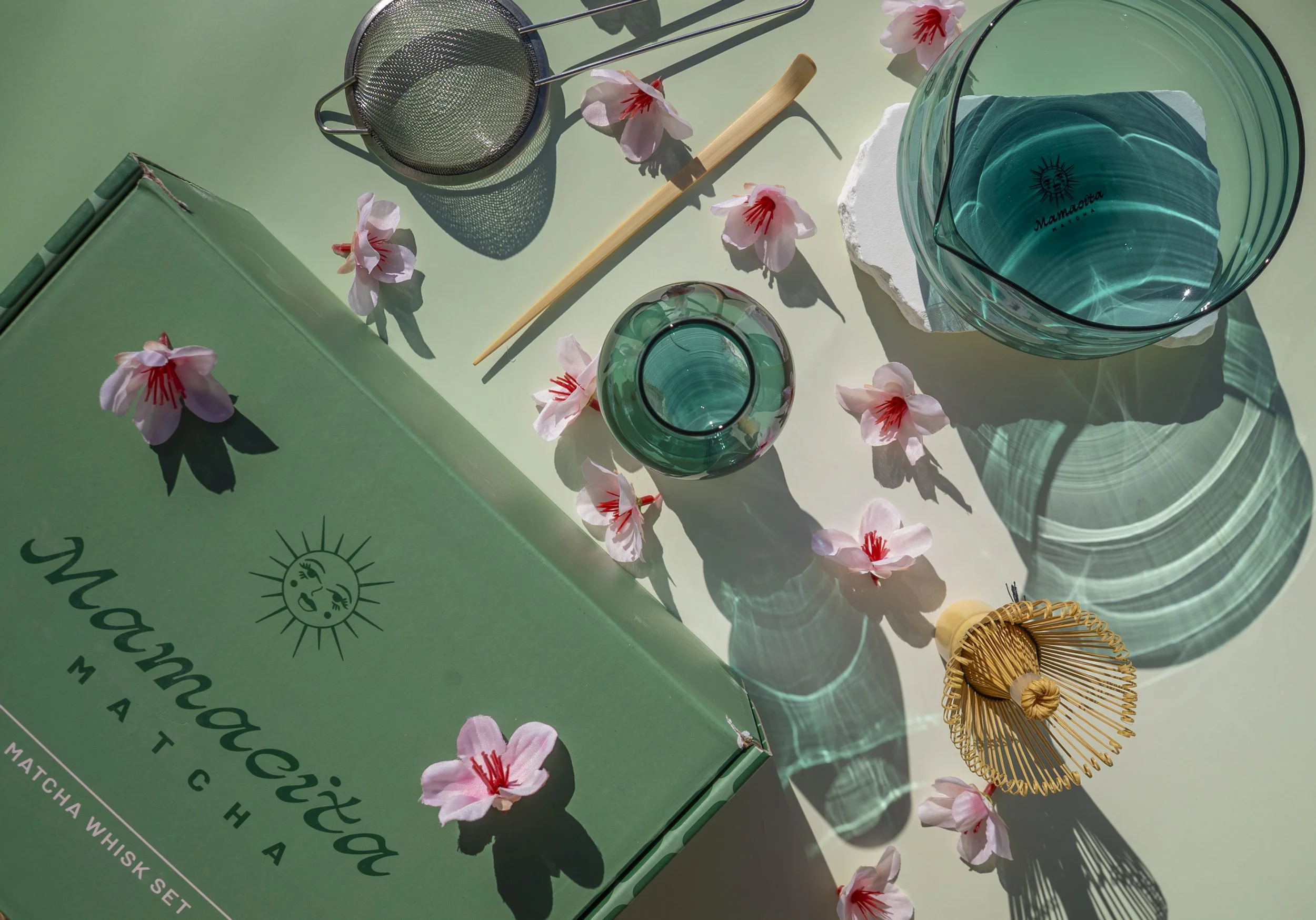A green box labeled Mamontoux Matcha, empty teal-colored glass bowls, a small teal vase, a small wooden whisk, pink flower petals, and a bamboo tea whisk, set on a light green surface with shadows and reflections from the glass items.