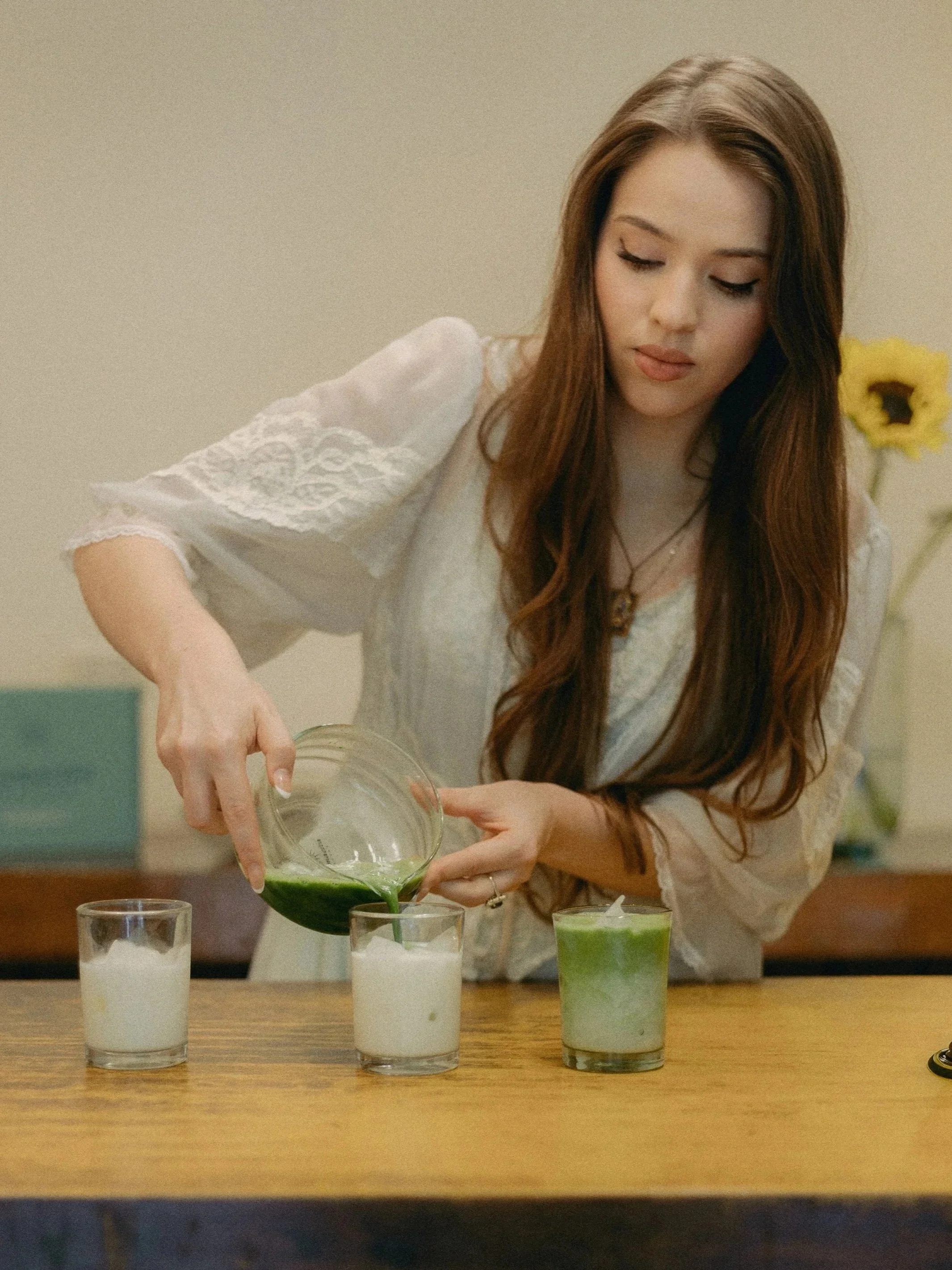 A young woman with long brown hair pouring a green beverage into glass with white liquid on wooden table, with yellow flowers in background.