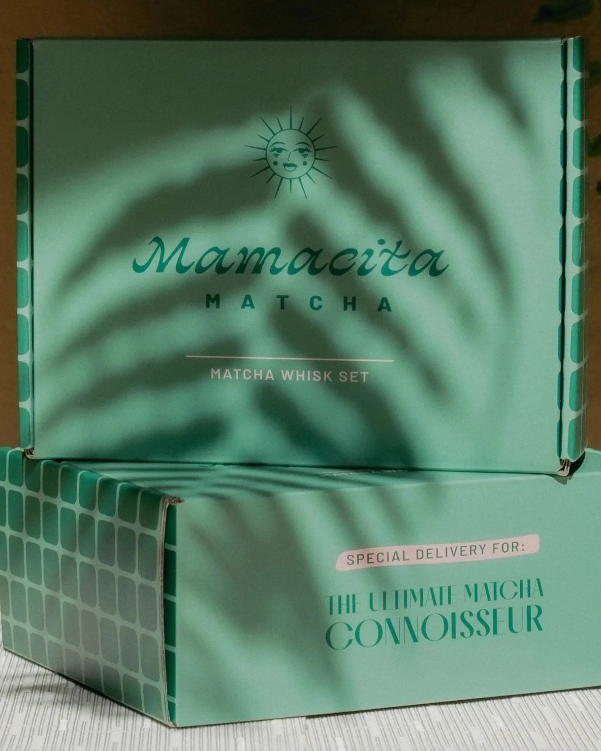 A light green box labeled 'Mamaçita Matcha' with a sun with a face logo. The box contains a matcha whisky set and is marked as 'special delivery for: the ultimate matcha connoisseur. Shadows from nearby leaves are cast on the box.