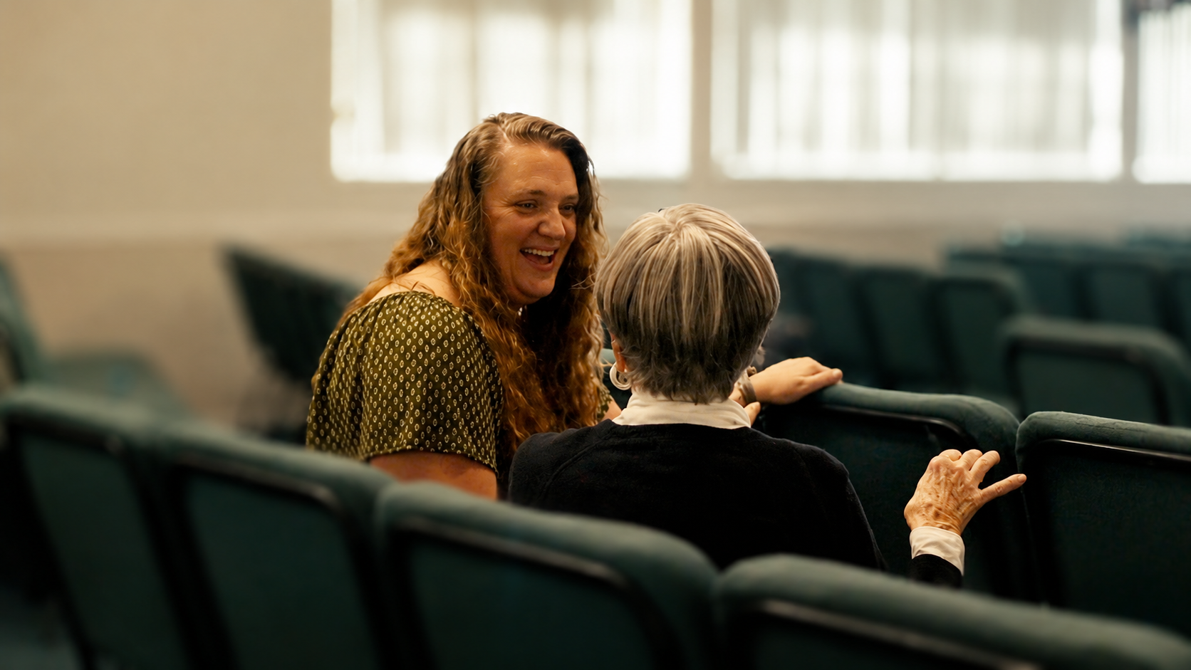 Two women, one young with long wavy hair and one older with short gray hair, are sitting in a nearly empty room with black chairs arranged in rows, having a conversation and smiling at each other.