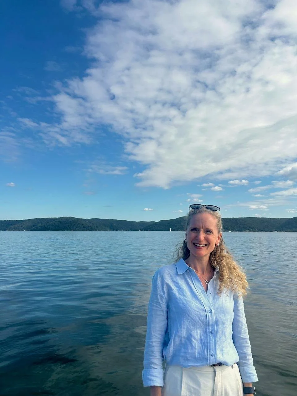 A woman with curly blonde hair, wearing a light blue shirt and white pants, standing by a large body of water with hills in the background and a partly cloudy sky.