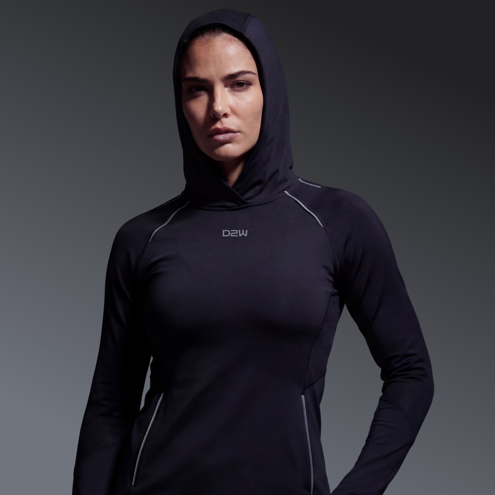 Woman in black athletic hoodie with hood up, posing with arms by her sides against a dark gradient background.