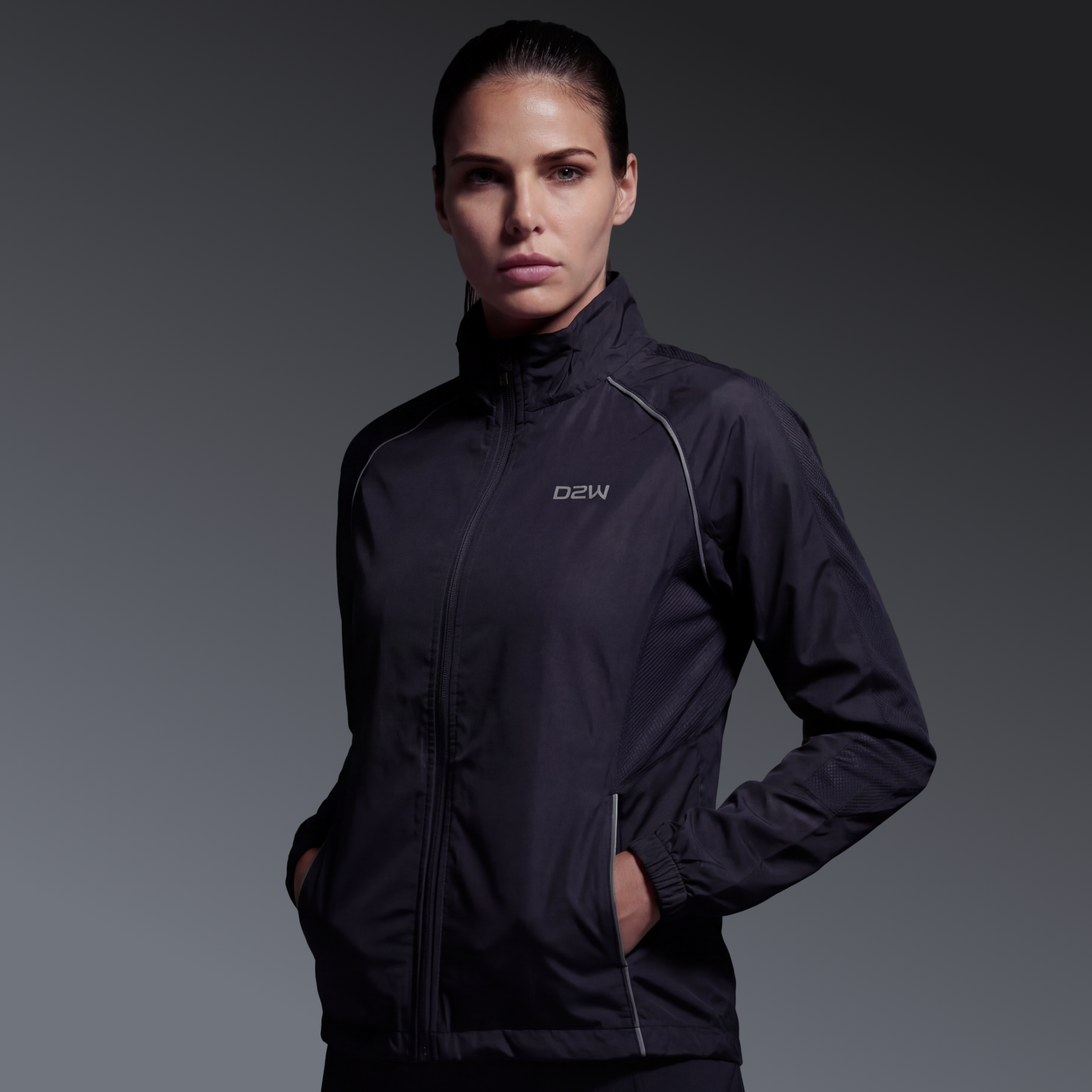 A woman wearing a black athletic jacket with her hands in the pockets, standing against a dark gray background.