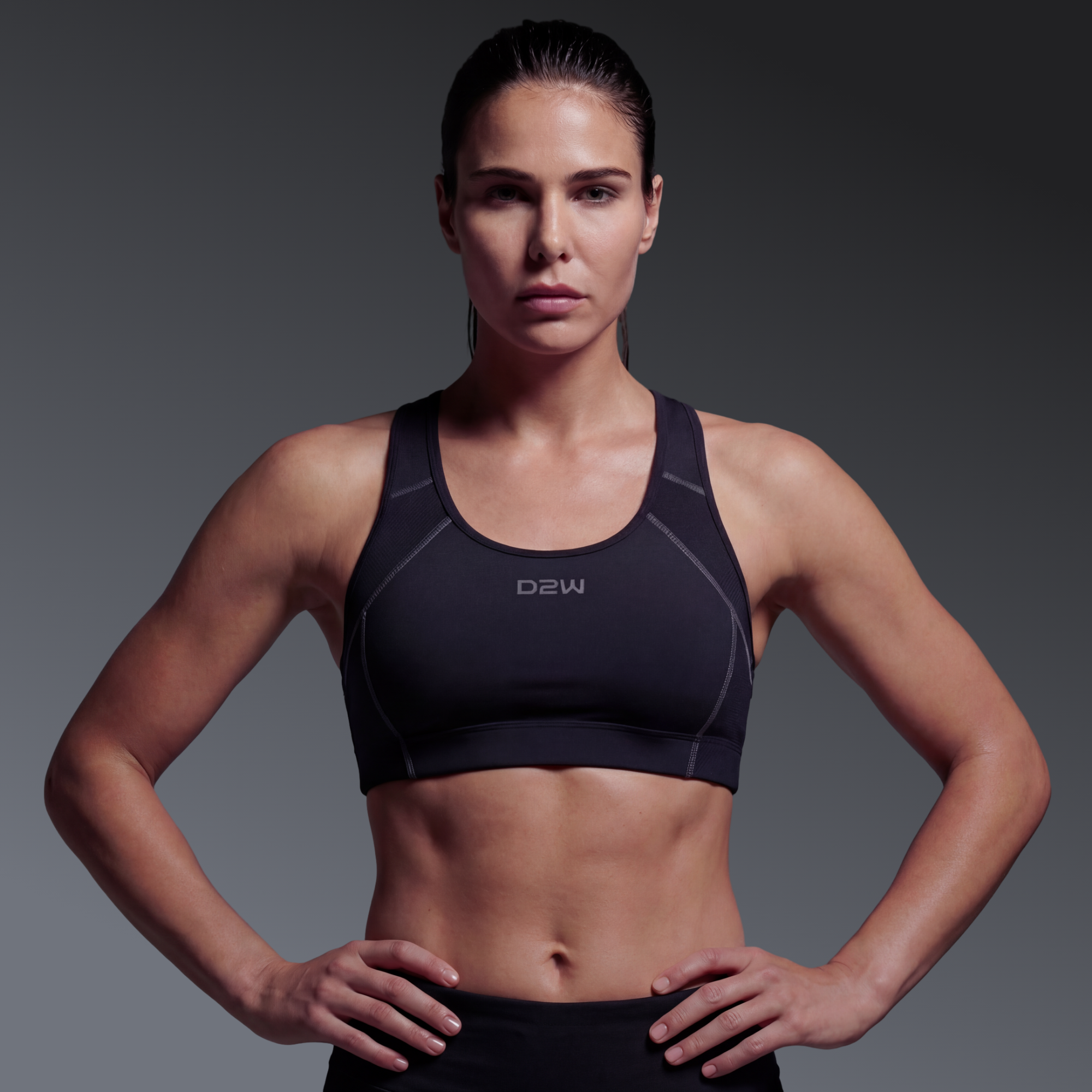 A fit woman with brown hair tied back, wearing a black sports bra, standing with her hands on her hips against a gray background.