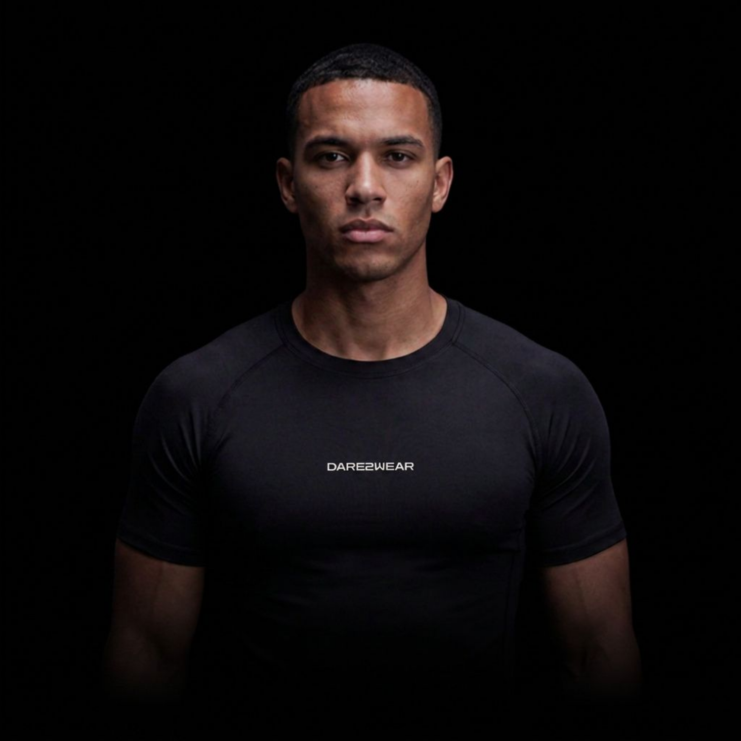 A young man with short dark hair, wearing a black athletic shirt with the word 'DARE2WEAR' printed on it, standing against a black background.