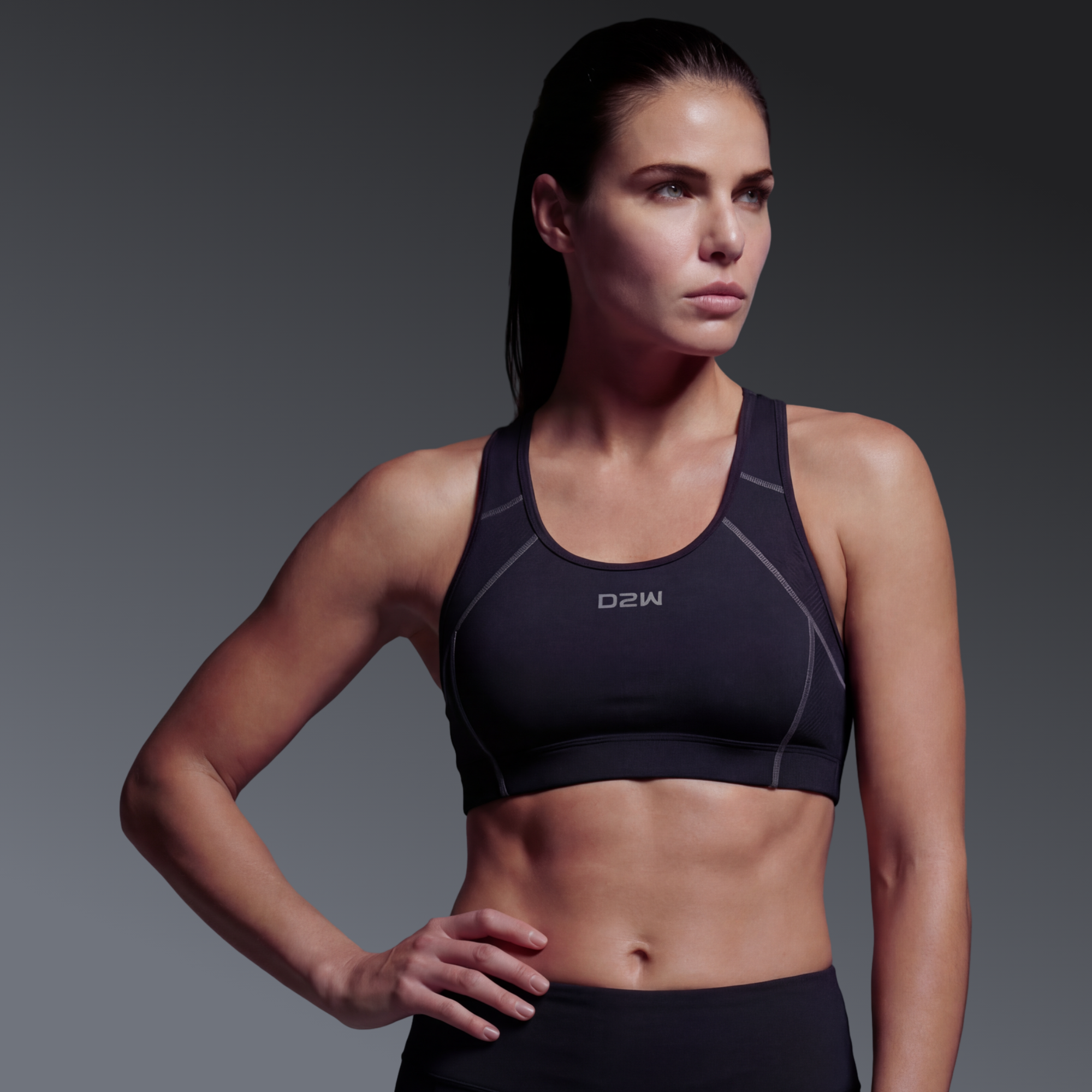 A fit woman in a black sports bra with gray accents and black leggings poses with one hand on her hip against a gray gradient background