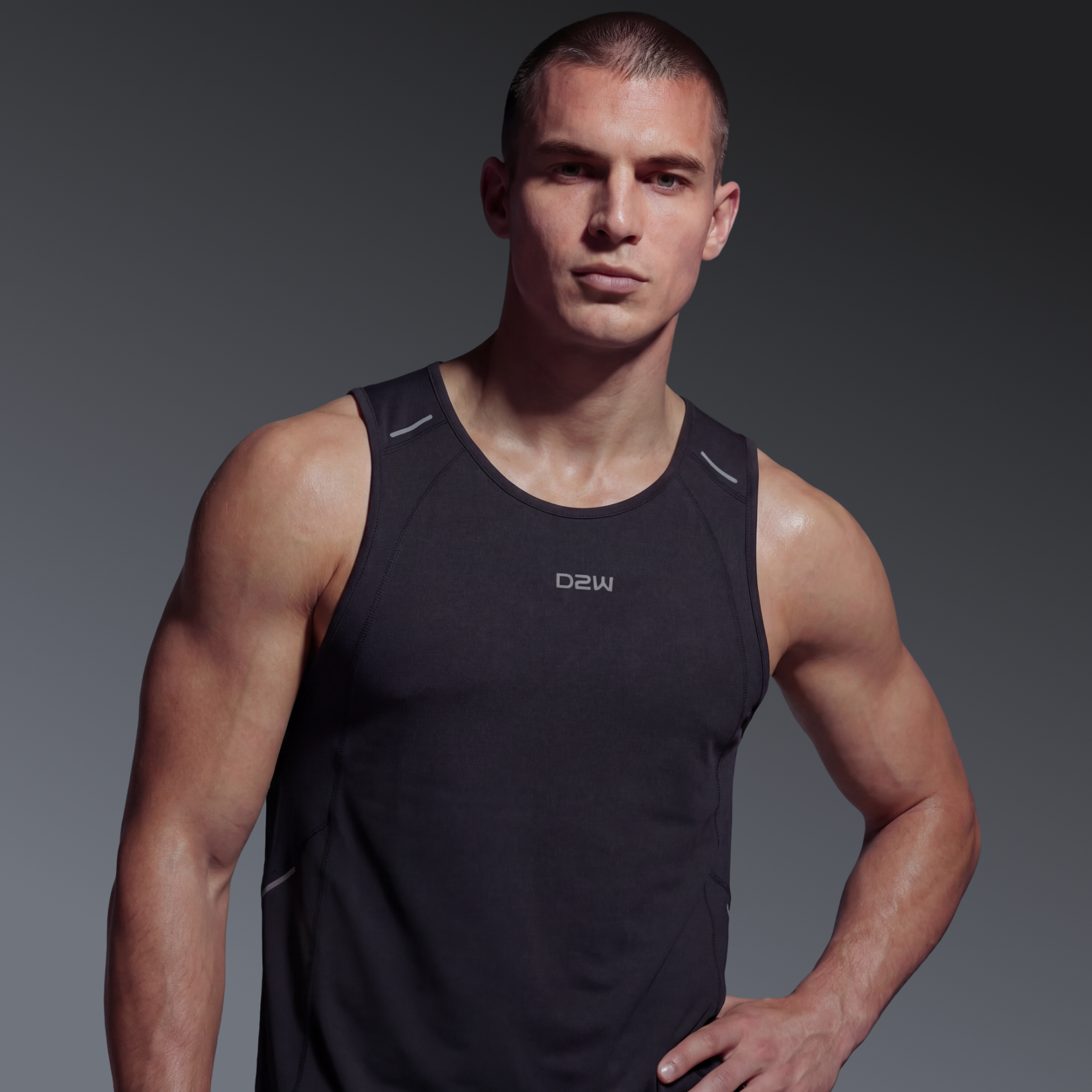 A muscular young man in a black athletic tank top stands confidently against a plain gray background.