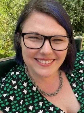 Headshot of Rachel Friendly, a white, cisgender woman wearing glasses, a silver necklace, and a green, black, and white patterned dress.