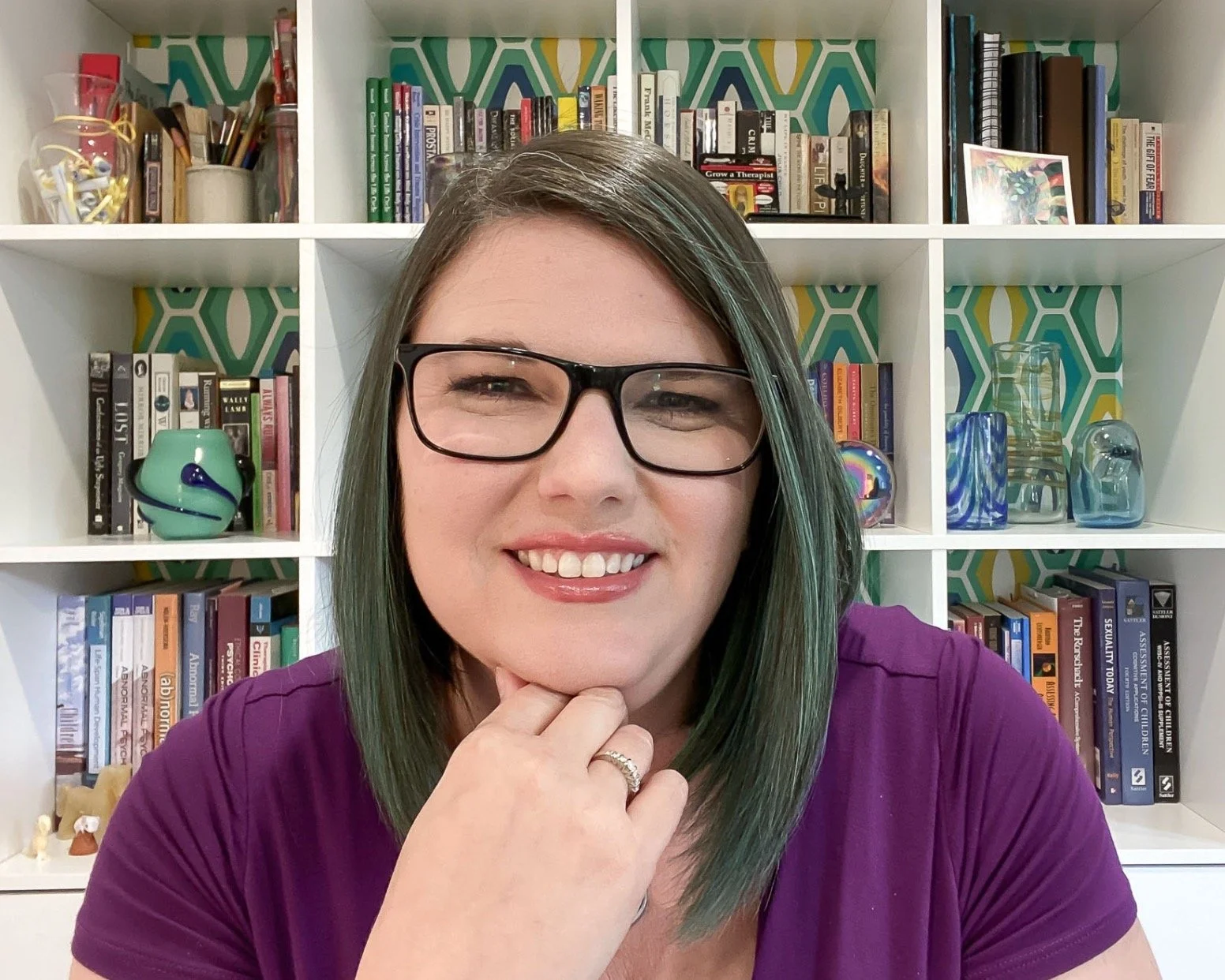 Rachel W. Friendly, Ph.D. is a Therapist in Fairfax, VA with straight dark hair streaked with aqua, a purple dress, and glasses. She is sitting with her chin on her hand, smiling.