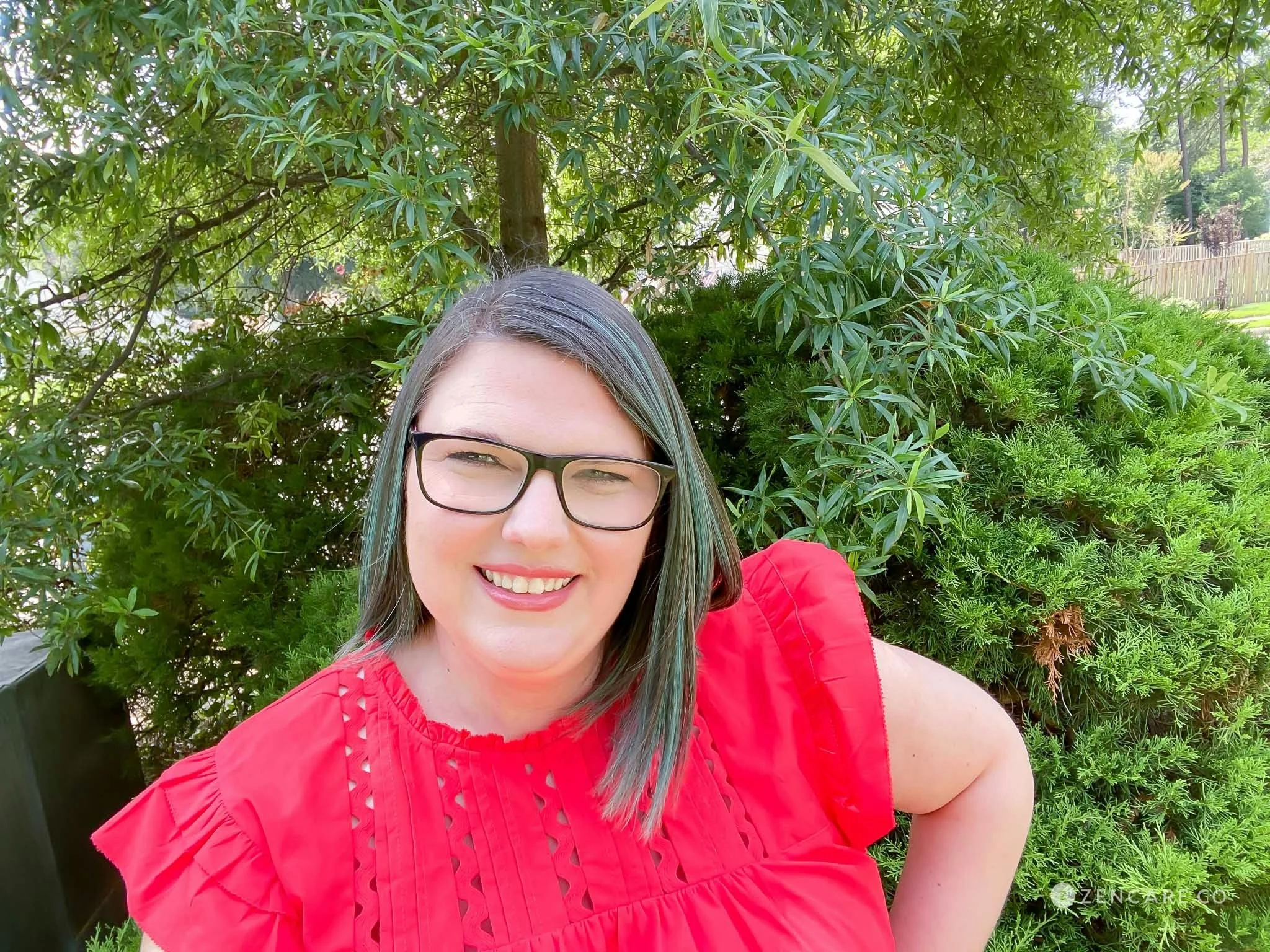 Rachel W. Friendly, Ph.D., Psychologist in Fairfax, VA, has pale skin and shoulder-length, straight, dark brown hair streaked with aqua. She is wearing a bright red top and glasses and standing in front of a green tree.