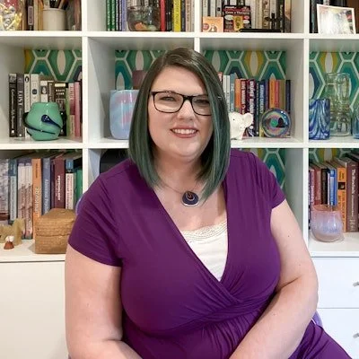 Rachel W. Friendly, PhD, Psychologist in Fairfax, VA has pale skin and dark brown, shoulder-length hair streaked with aqua. She is wearing a purple dress, a necklace, and glasses and standing in front of a colorful bookcase.