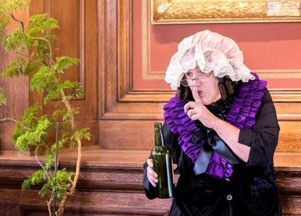 A woman dressed in historical costume, wearing a large bonnet, purple ruffled shawl, and black dress, standing in a wood-paneled room. She is holding a green bottle and making a shushing gesture with her finger to her lips.