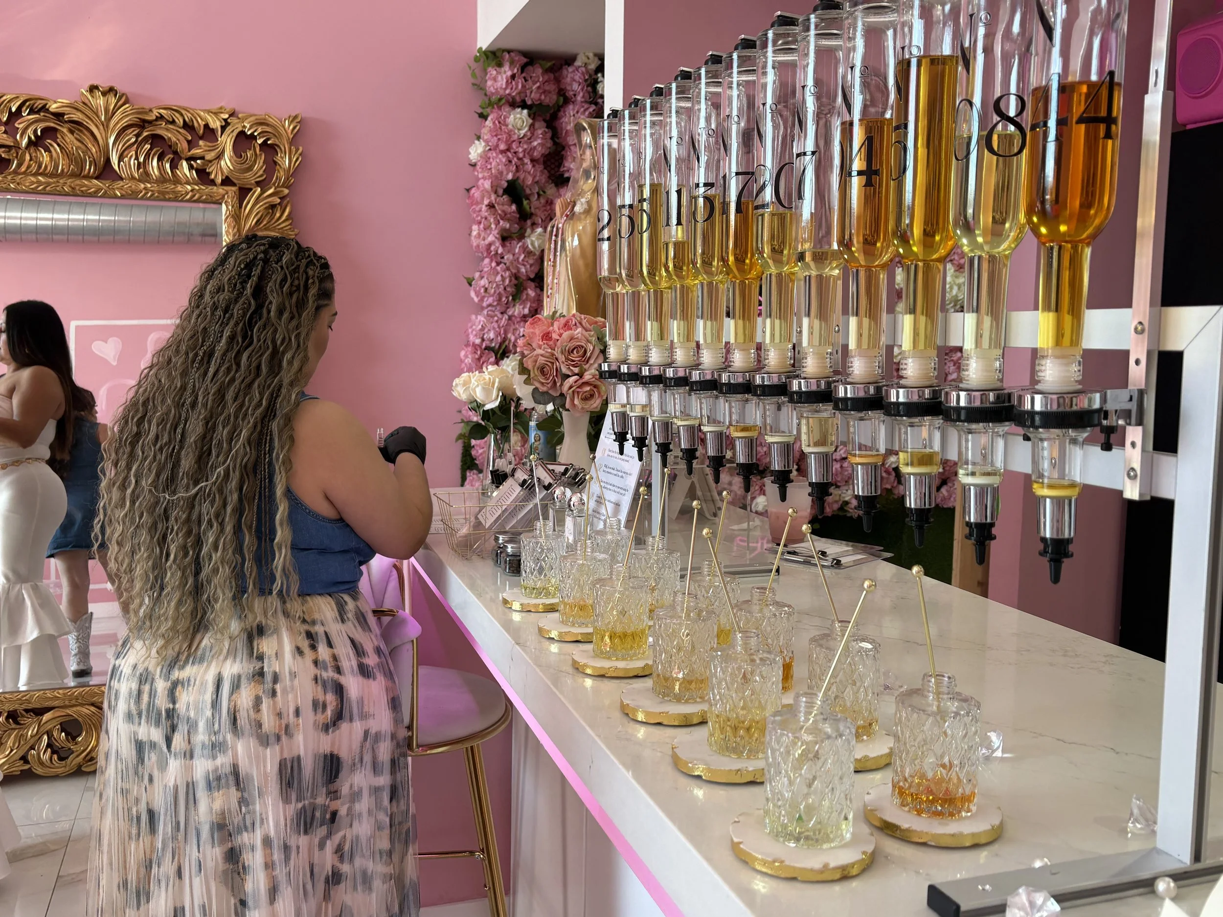 A woman with long curly hair in a blue top and leopard print skirt standing at a bar counter with multiple dispensers filled with colored liquids, serving drinks in ornate glasses with gold trim, in a pink-themed venue decorated with flowers and gold accents.