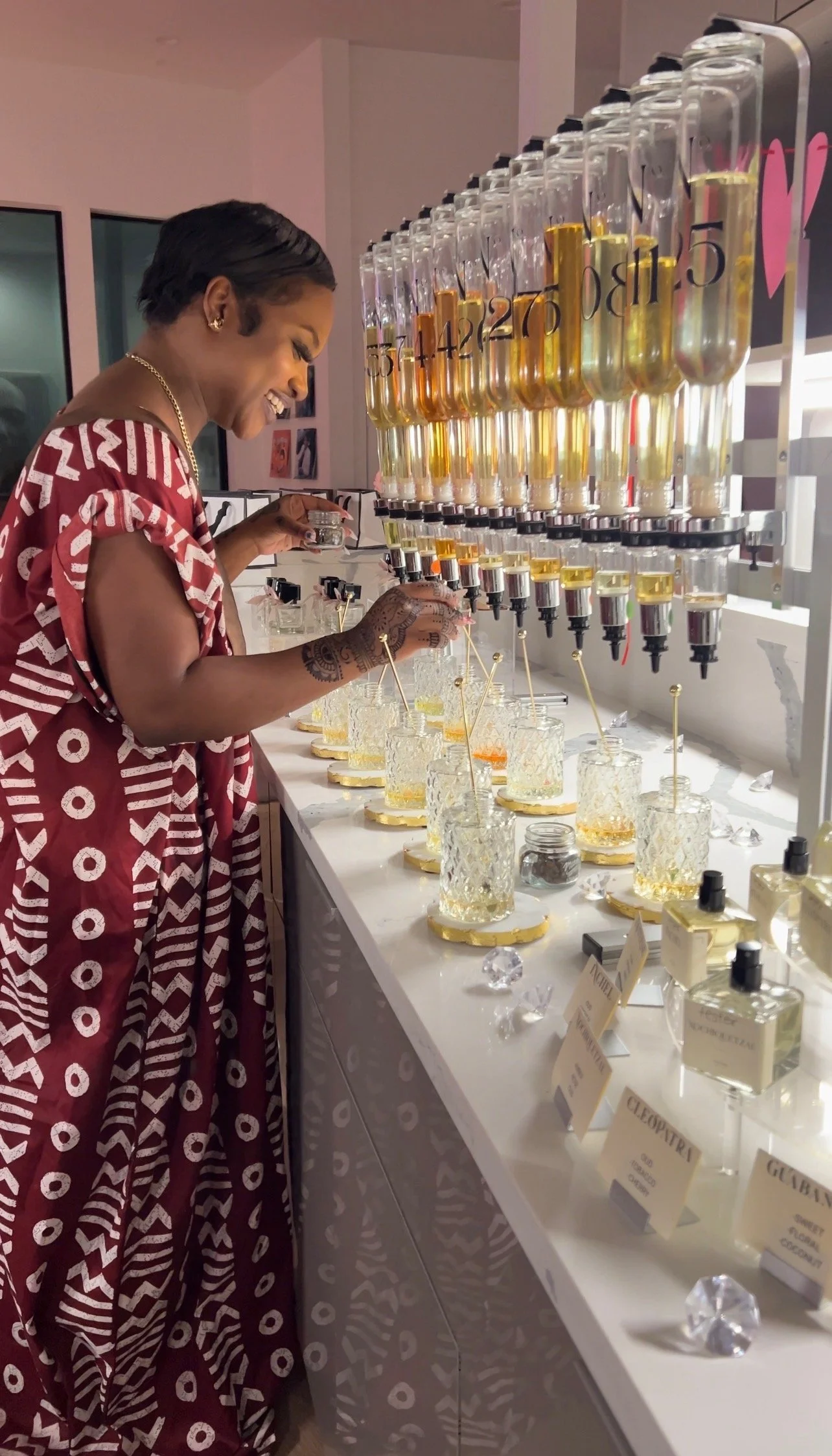A woman with short black hair, wearing a red and white patterned dress, smiles while preparing drinks at a counter with liquid dispensers and glassware, possibly in a bar or beauty salon.