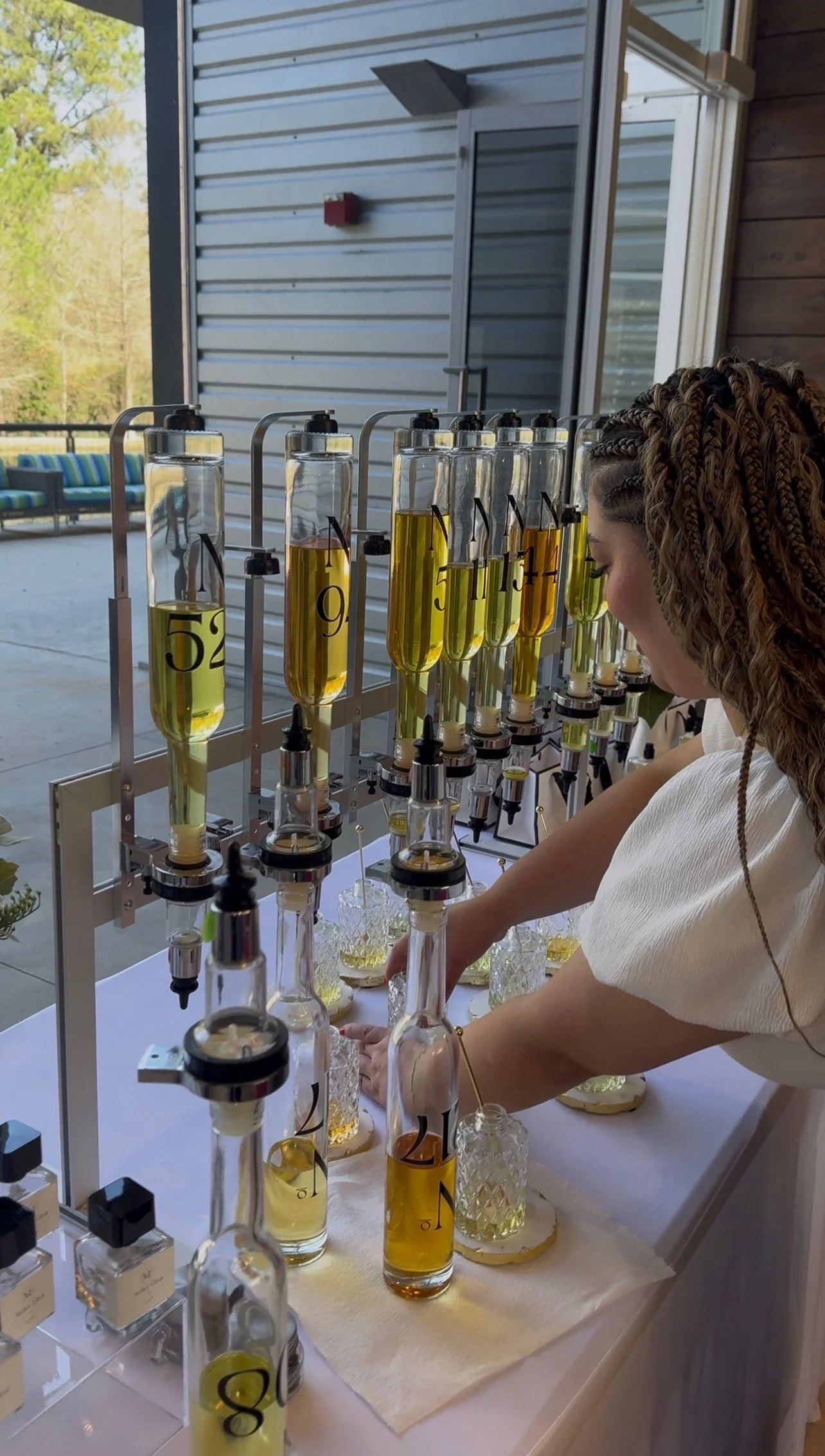 A woman is working at a table with multiple glass containers filled with yellow liquid, each labeled with a number, connected to equipment to distill or analyze the liquids. She is handling the setup with her hands, and the scene is set outdoors on a patio with a building wall and trees in the background.