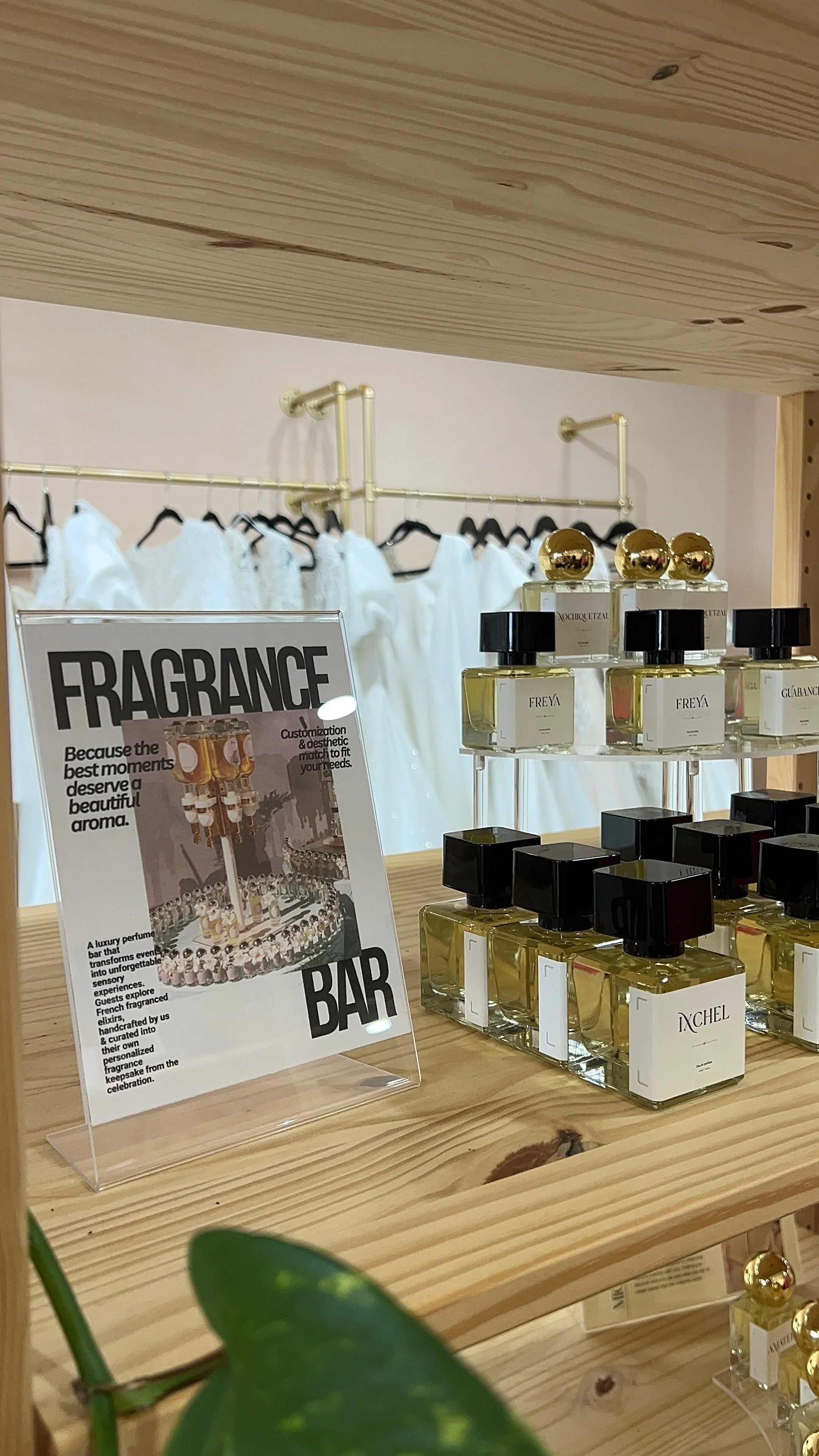 Display of perfume bottles on a wooden shelf with a framed sign that reads 'Fragrance Bar' and a clothing rack with white garments in the background.