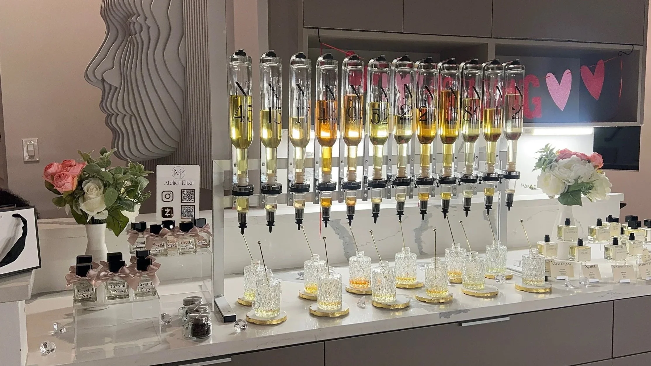Display of perfume testing stations with glass bottles filled with various scents, decorative flowers, and a sign for Atelier Elixir.