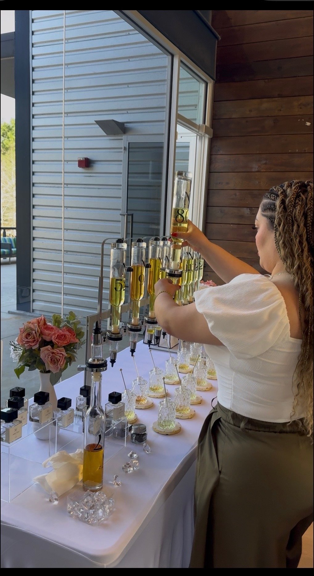 A woman makes cocktails at a bar with multiple infusion tubes filled with yellow liquid, bottles of ingredients, and decorative flowers on the table.