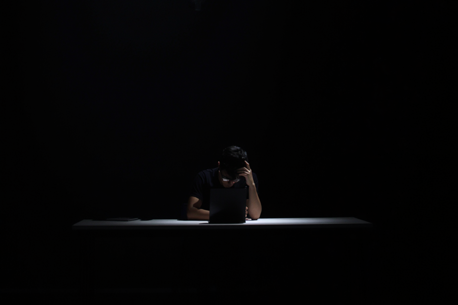 A person sitting alone at a white table in a dark room, working on a laptop with a focused or stressed expression, hand on their forehead.