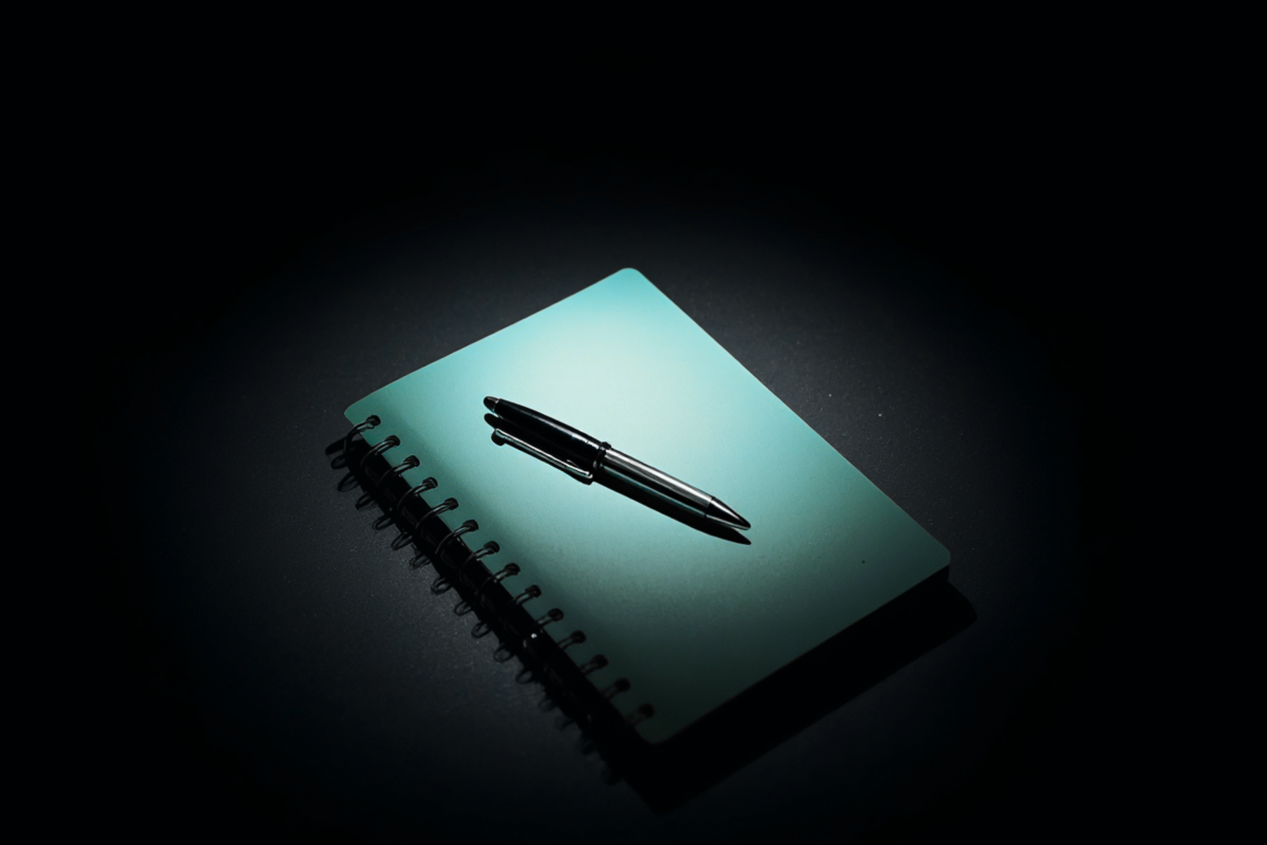 A spiral-bound notebook with a black pen resting on top, illuminated against a dark background.