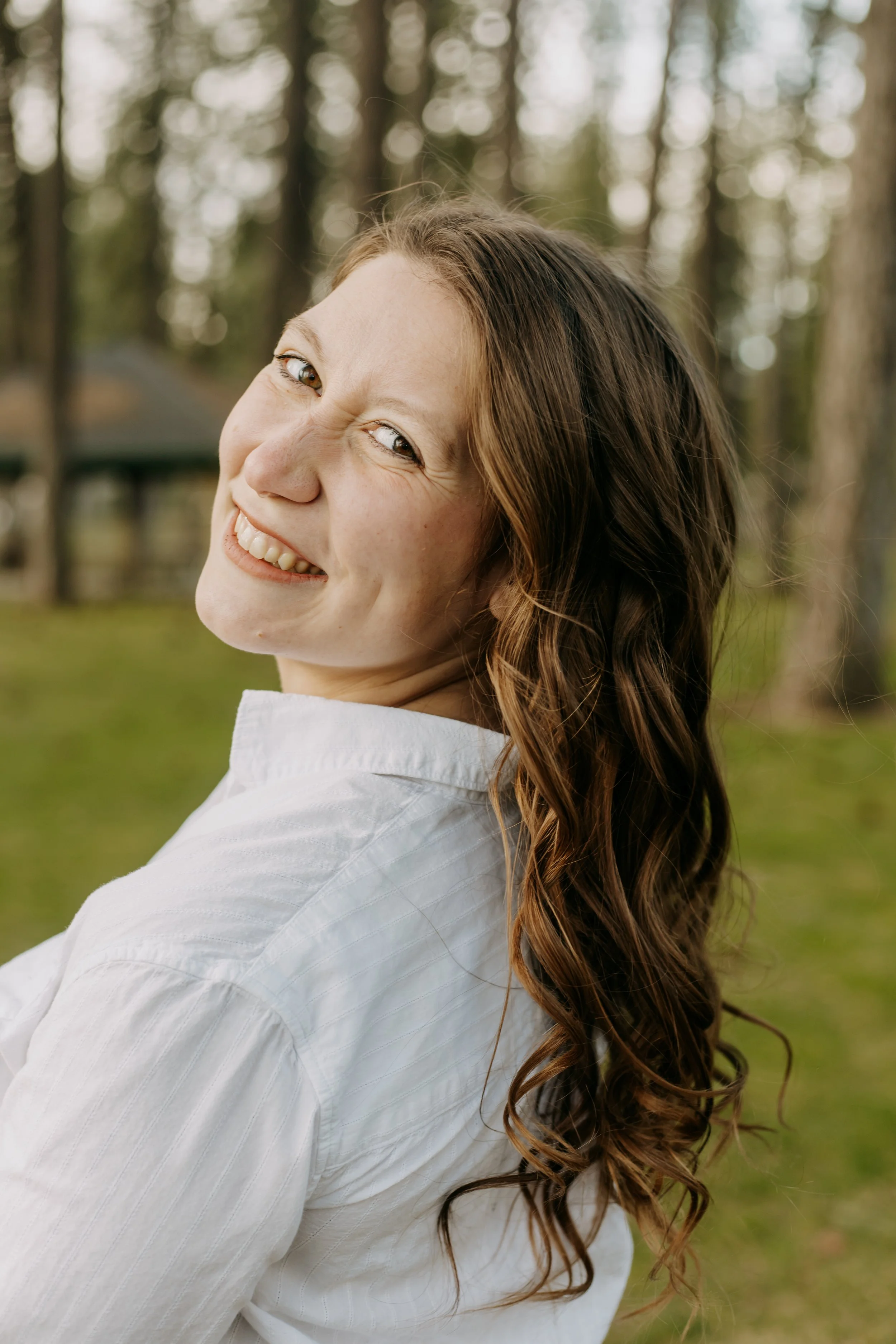 A smiling woman with long, wavy brown hair, wearing a white shirt, outdoors in a wooded area.