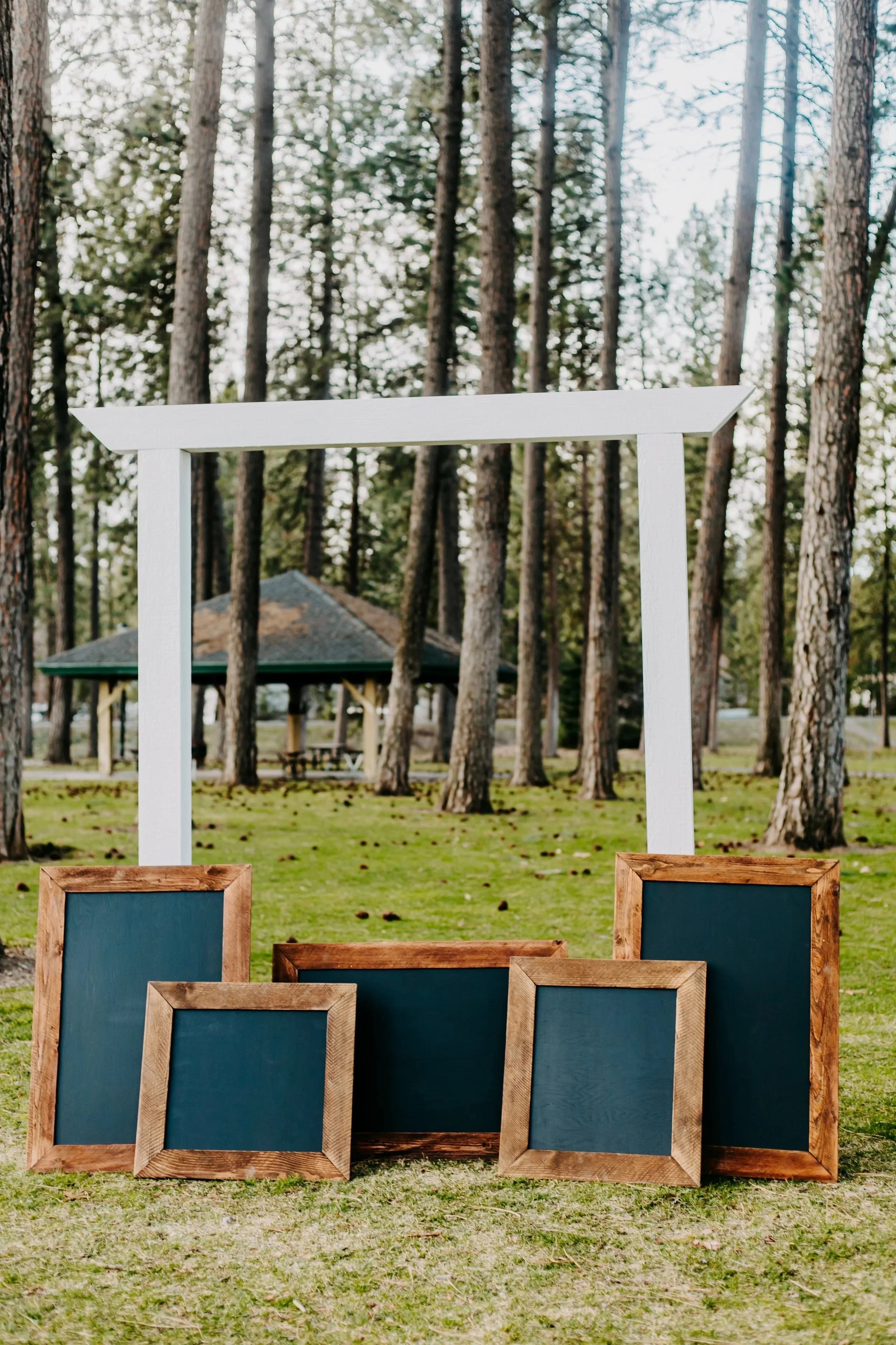 Rustic wood framed chalkboards leaning against a white square arch in a park with trees and a pavilion in the background.