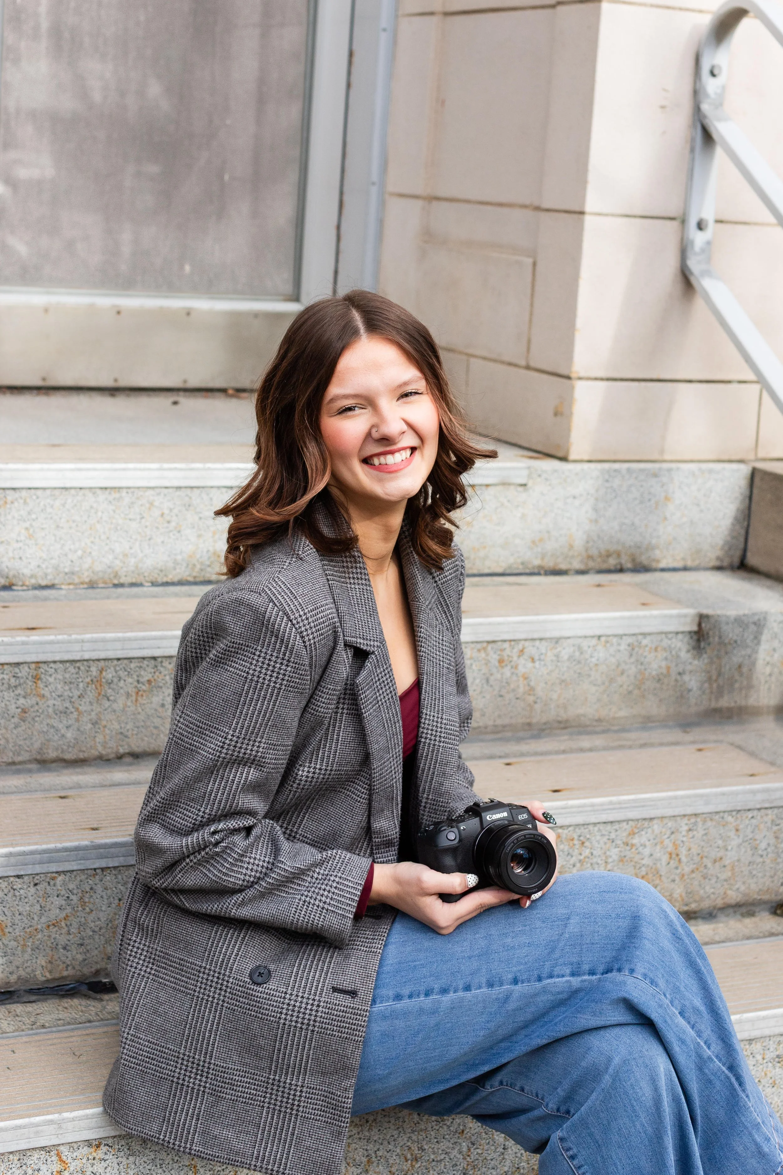 A young woman with brown hair sitting on outdoor stairs, smiling and holding a camera.