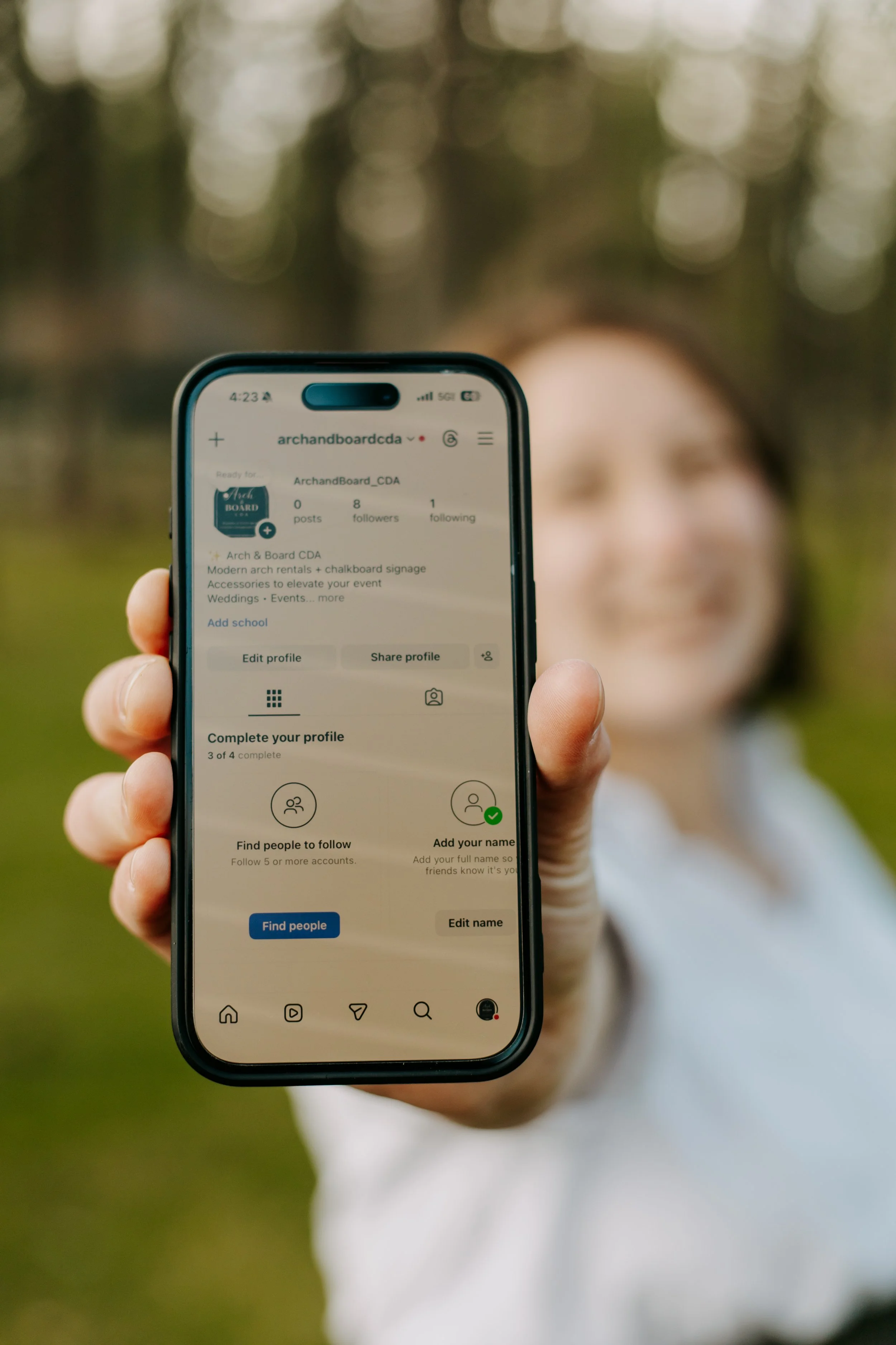 Person holding a smartphone displaying an Instagram profile with the username 'archandboardcda' against a blurred outdoor background.