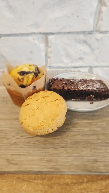 Three baked goods on a wooden surface in front of a white brick wall. A muffin with chocolate chips in paper wrapper, a yellow cookie, and a chocolate glazed donut with powdered sugar.