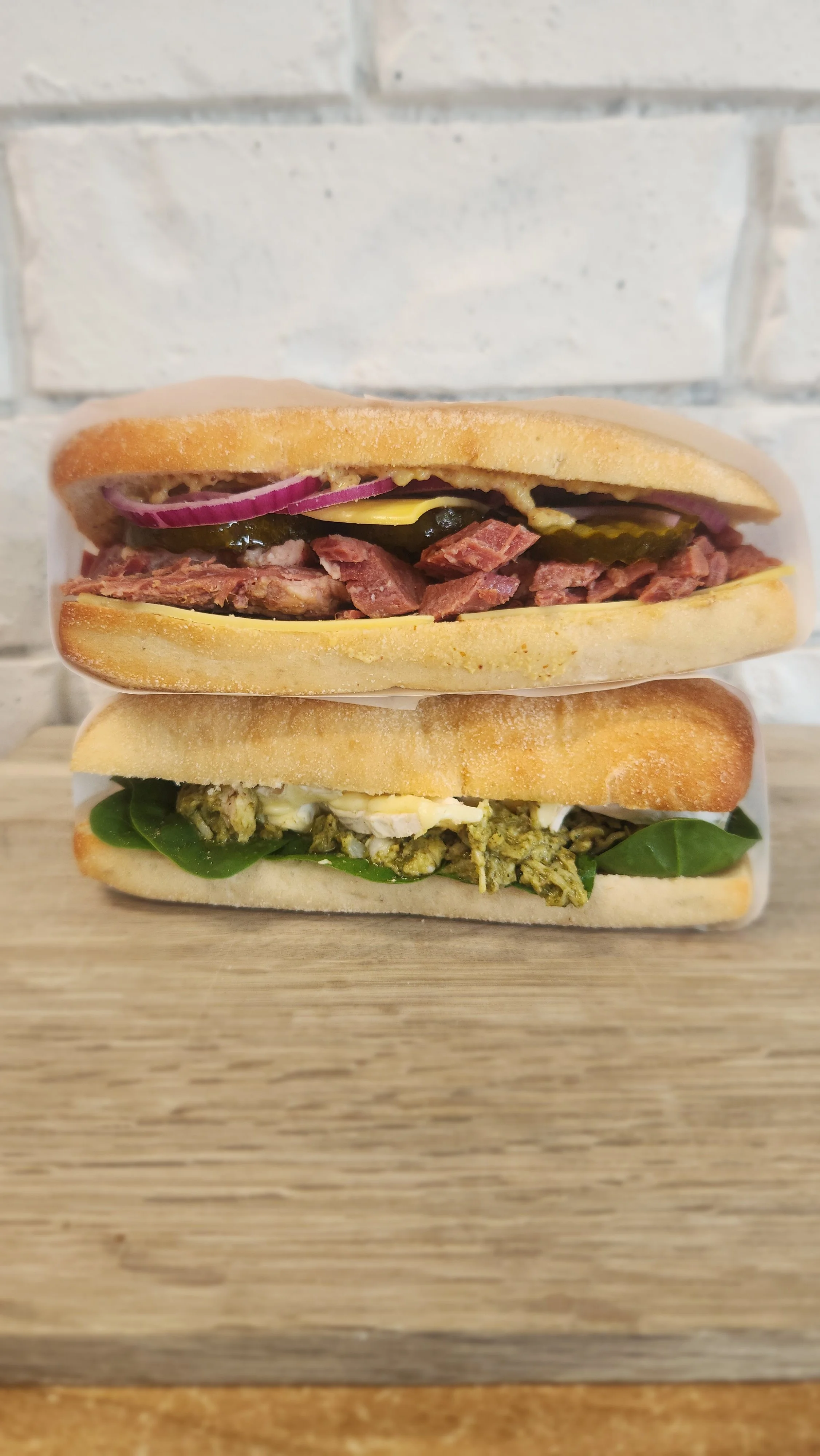 Two sandwiches stacked on a wooden surface with a white brick wall in the background.