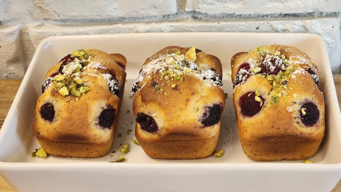 Three blueberry muffins topped with chopped pistachios and powdered sugar in a white dish on a wooden surface with a brick wall background.