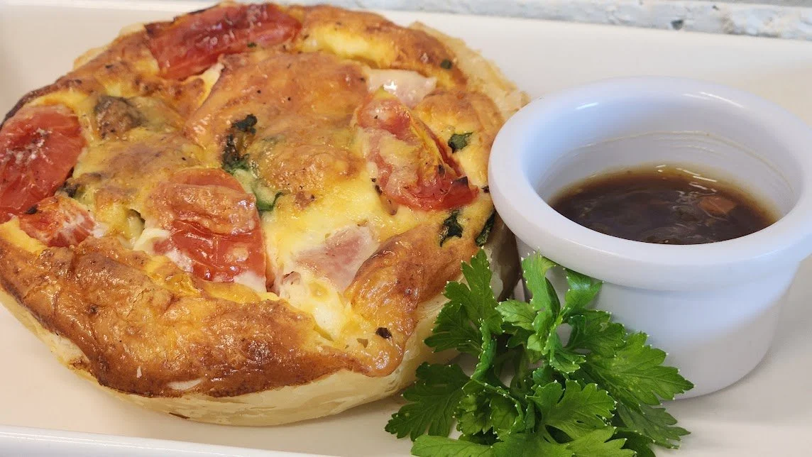 A savory breakfast biscuit sandwich with melted cheese, pepperoni, and vegetables, served with a side of dark soy sauce or gravy and garnished with fresh parsley.