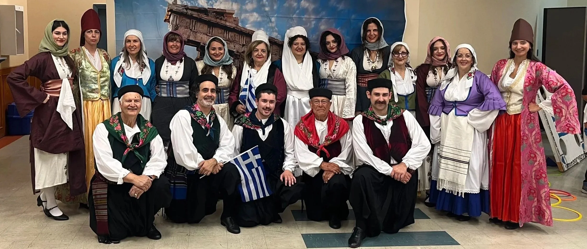 Group of people dressed in traditional Greek costumes, some women wearing headscarves and long skirts, and men in vests and scarves, posing indoors with a backdrop of Greek ruins and flags.
