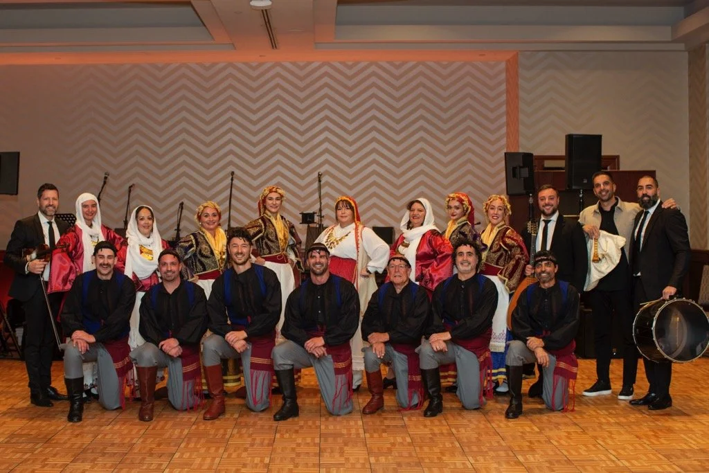 A group of people dressed in traditional Greek costumes, some women in embroidered dresses and headscarves, and men in black shirts and striped pants, some holding musical instruments, in an indoor hall with a patterned wall and a wooden floor.