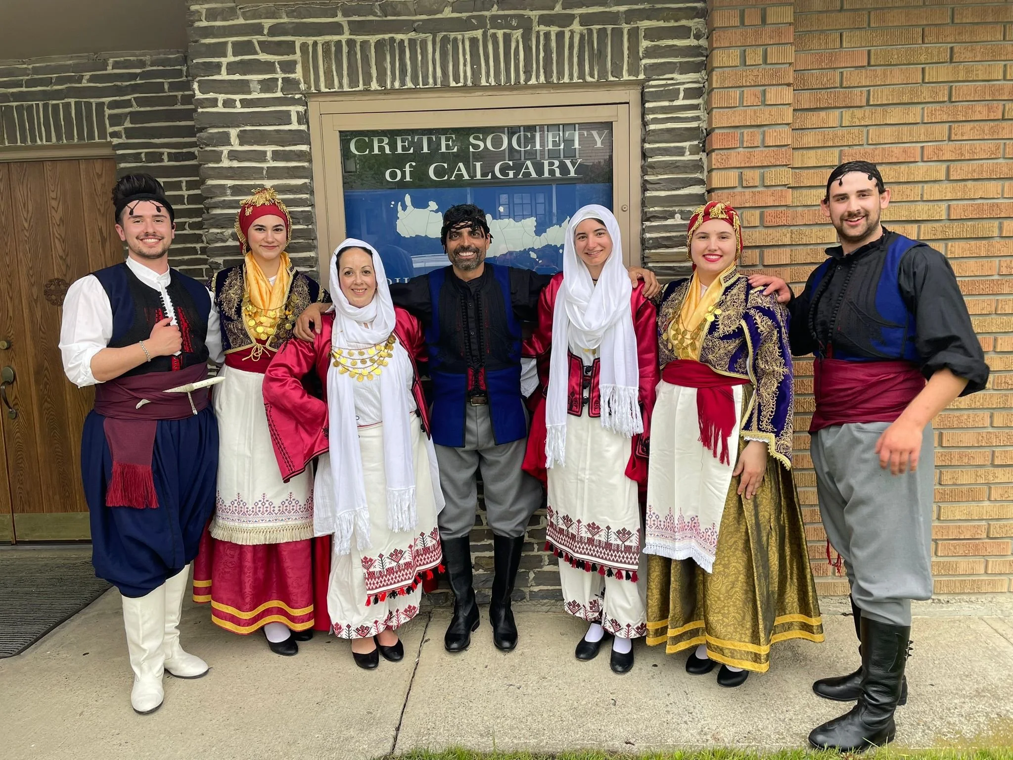 Group of seven people in traditional Greek attire standing outside in front of a building with a sign that reads "Crete Society of Calgary". The group is smiling, with three men and four women, all dressed in colorful, elaborately decorated costumes.
