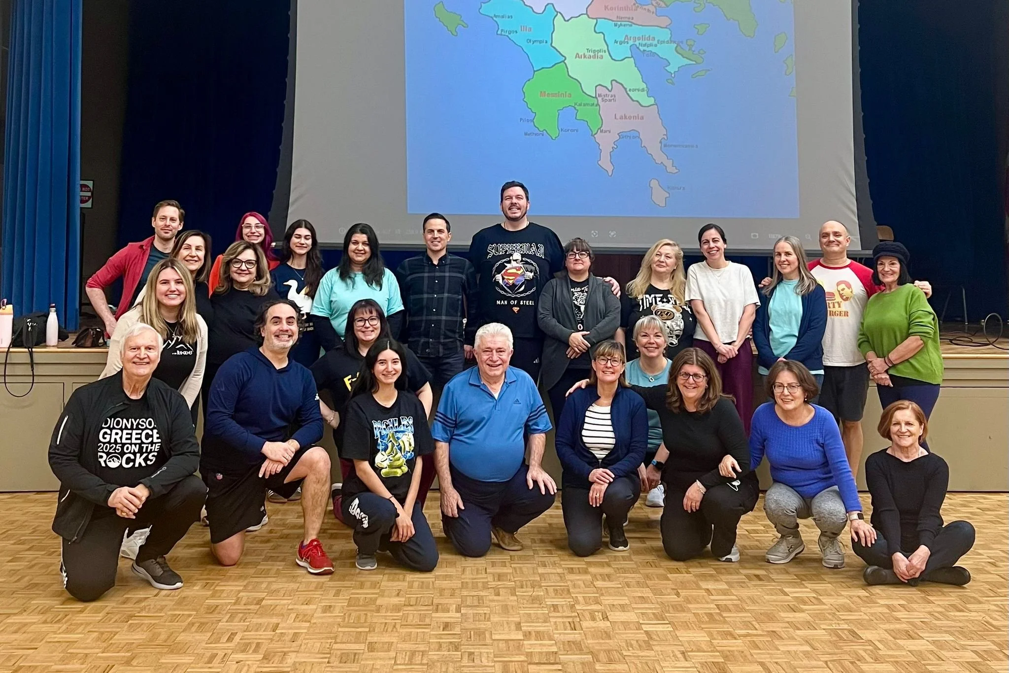 Group of people posing for a photo in a hall with a large map of Greece projected on the wall behind them.