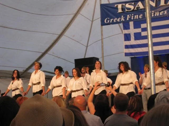 Group of women dressed in white blouses and black belts performing on stage at a Greek cultural event, with an audience watching.
