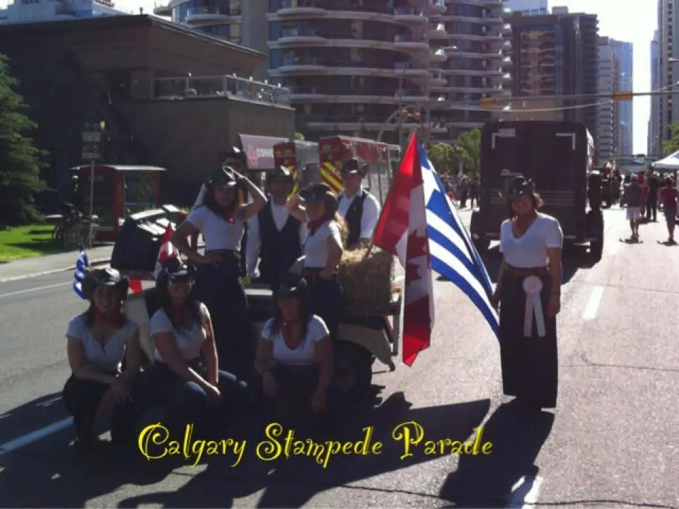 Group of women dressed in white shirts and dark skirts participating in the Calgary Stampede Parade, with a float decorated with hay and a Canadian flag in an urban street setting.