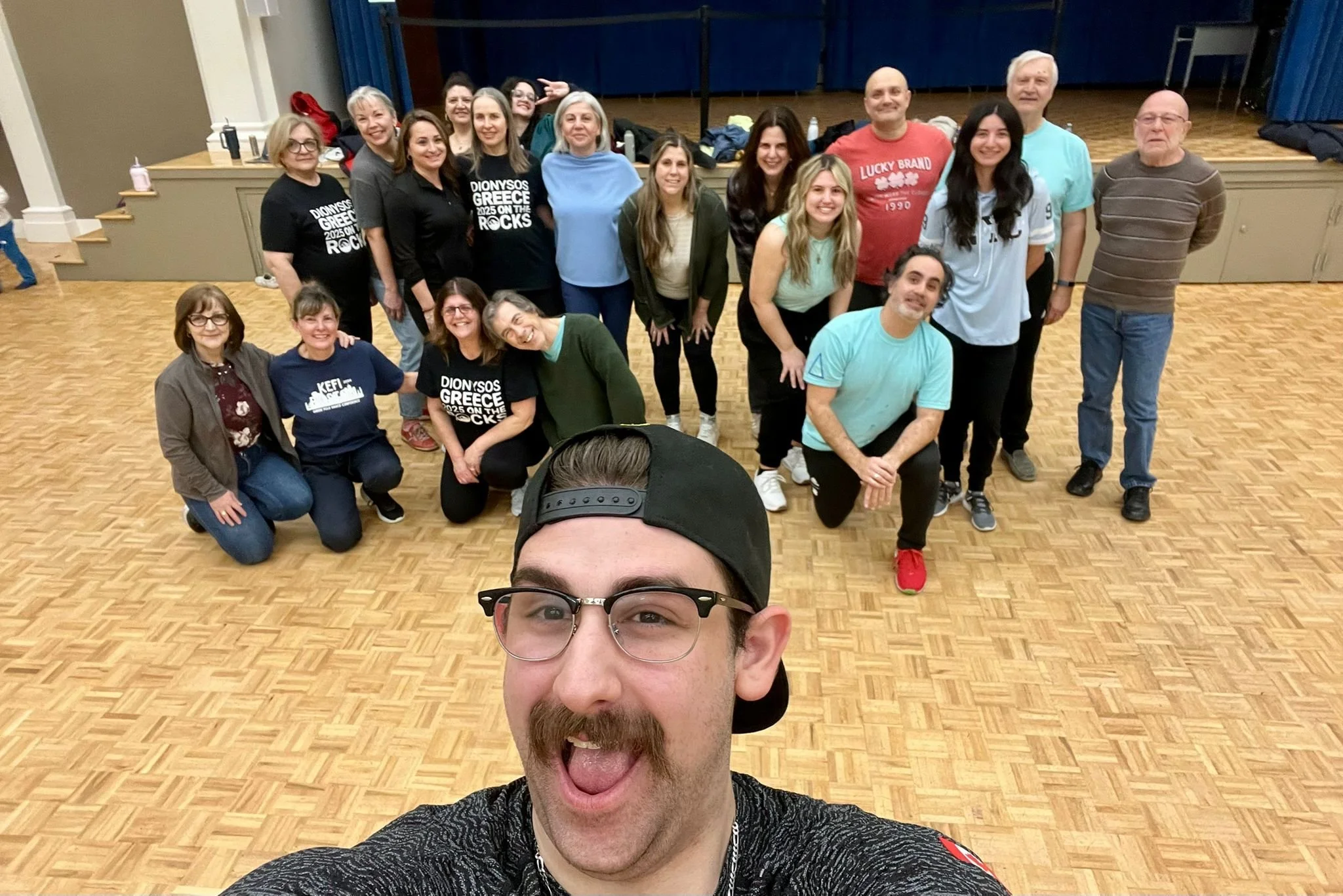 A group of about 15 people standing and smiling in a room with wooden flooring, with one person in the front taking a selfie and sticking out his tongue. The group includes men and women of various ages, some wearing casual clothing and others in T-shirts with text on them.
