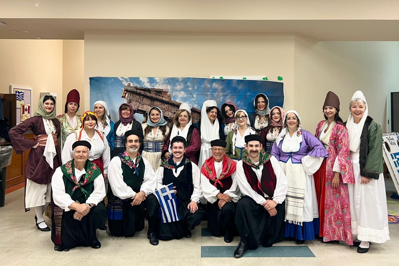Group of people dressed in traditional Greek costumes posed indoors in front of a backdrop depicting a Greek landscape with ruins and blue sky.