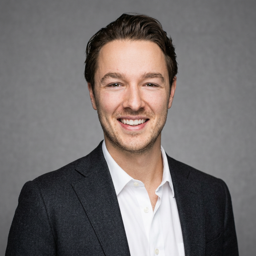 A smiling young man with short brown hair wearing a dark suit jacket and white shirt against a plain gray background.