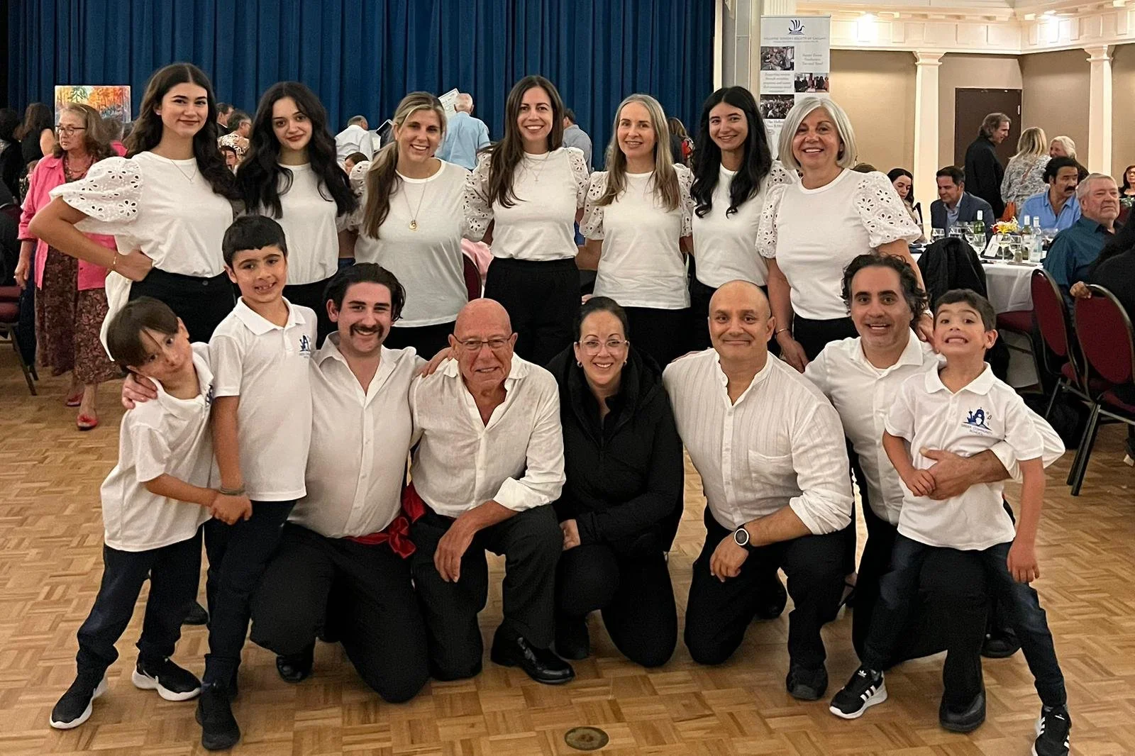 A group of 14 people, including children and adults, posing together at an indoor event with tables and a blue curtain in the background. They are smiling and wearing mostly white shirts.