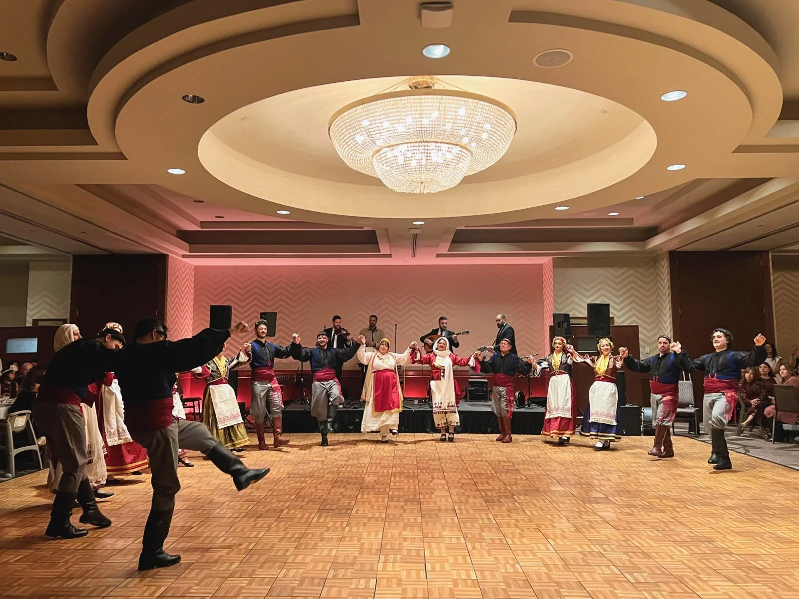 A group of people in traditional clothing performing a dance on a dance floor in a banquet hall with a large chandelier overhead.