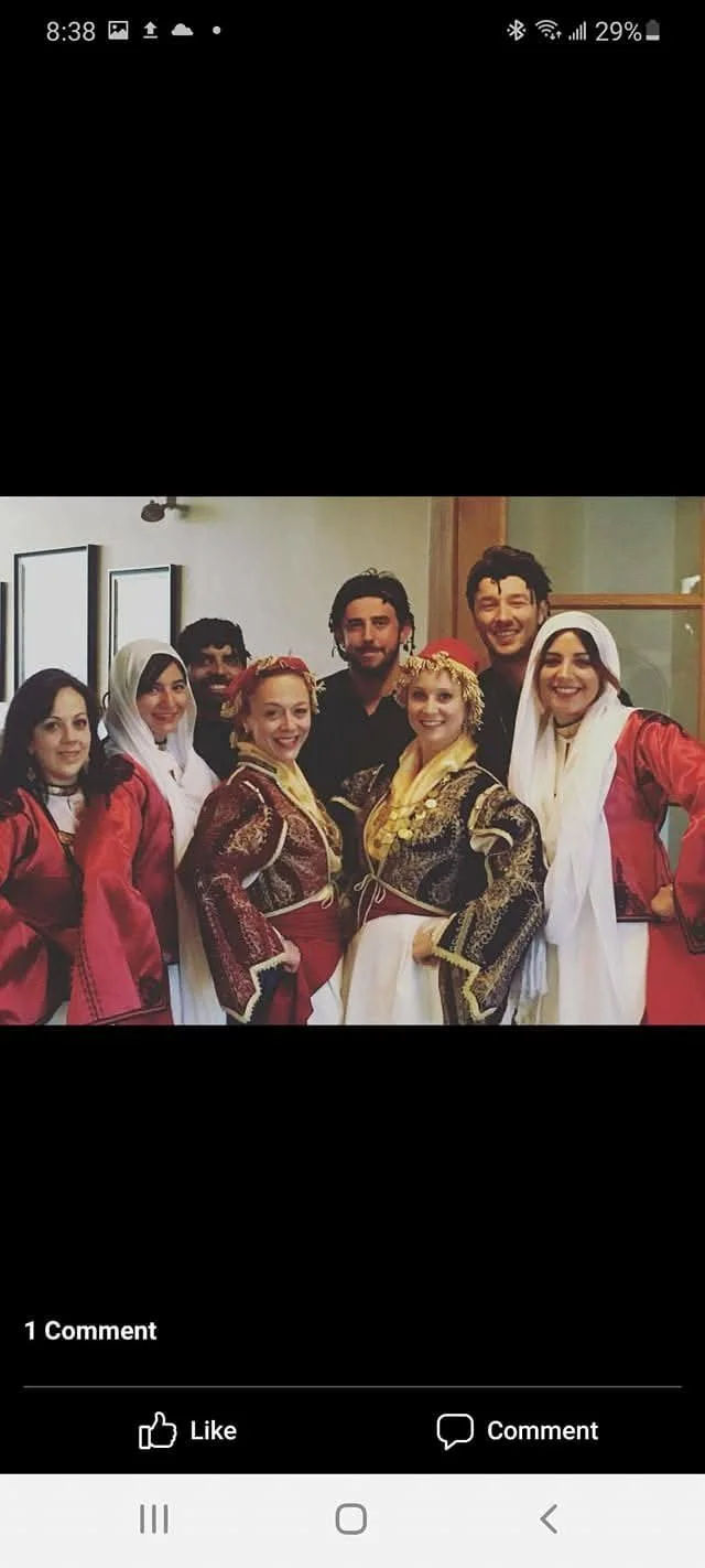 A group of seven people dressed in traditional Balkan or Mediterranean folk costumes, posing indoors and smiling at the camera.