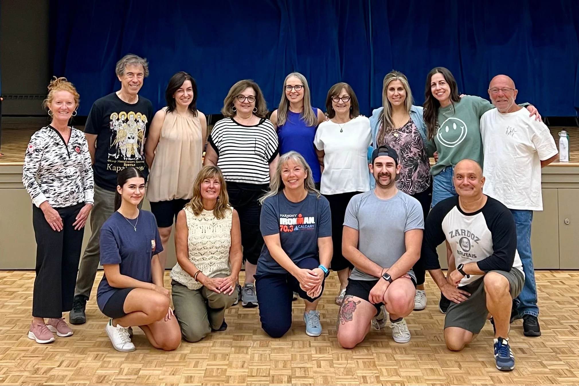Group of fourteen smiling people in casual clothes posing together in front of a blue curtain and wooden floor in an indoor setting.