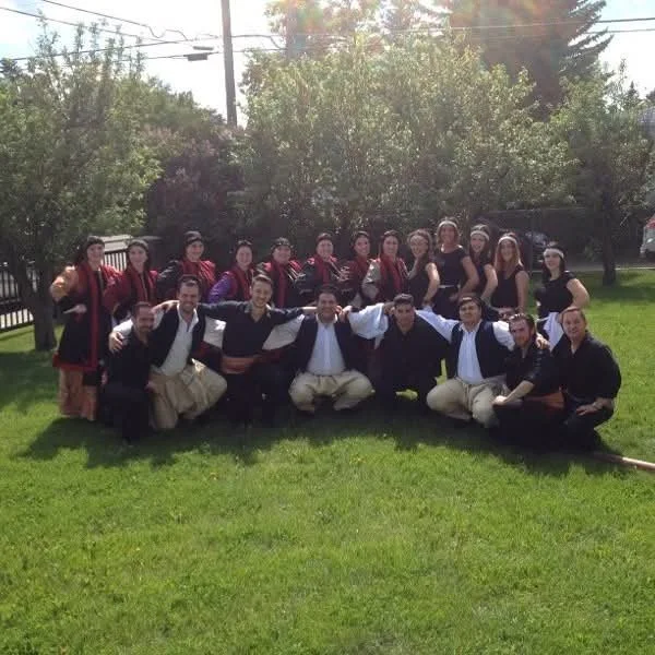 Group of people in traditional Eastern European clothing, posing outdoors on a grassy area with trees in the background.