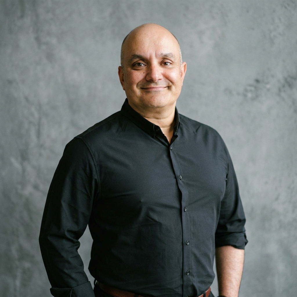 A man with a bald head smiling, wearing a black button-up shirt, standing against a gray textured background.